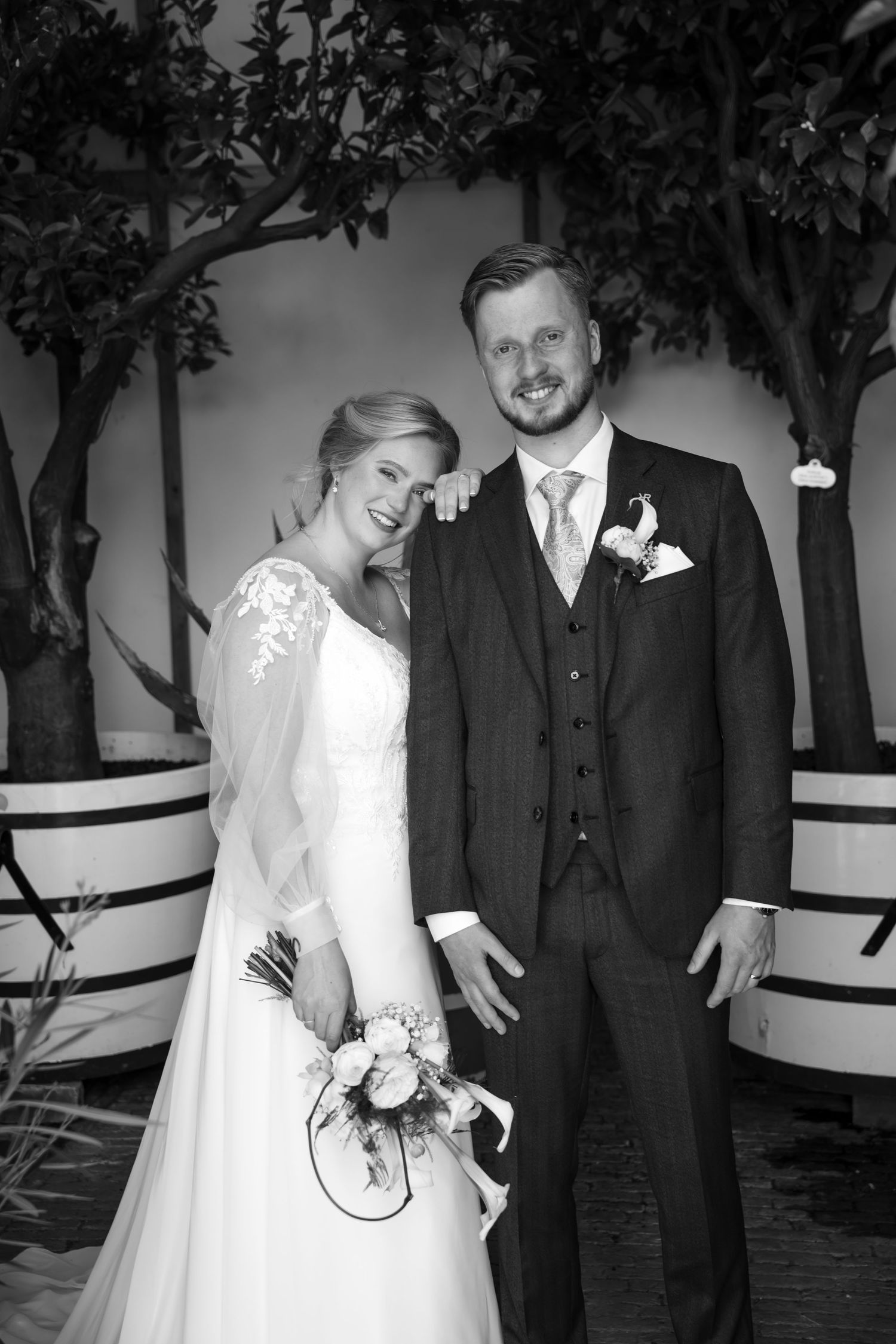 Black and white wedding portrait of smiling bride in lace gown holding bouquet with groom in suit among trees.