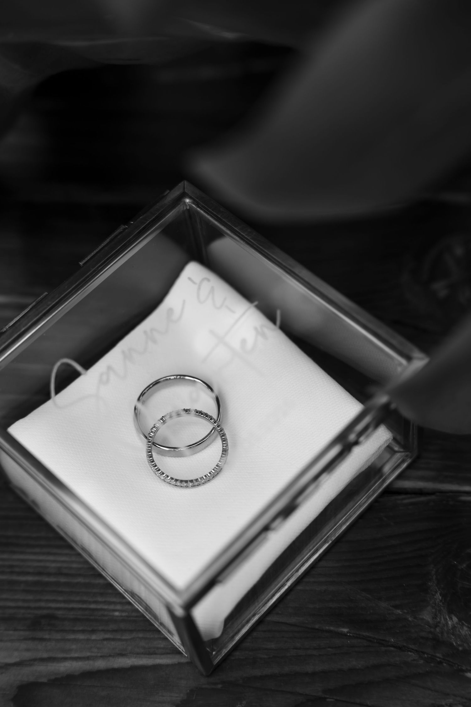 Two wedding bands rest on a white cushion inside a glass jewelry box, captured in elegant black and white photography.