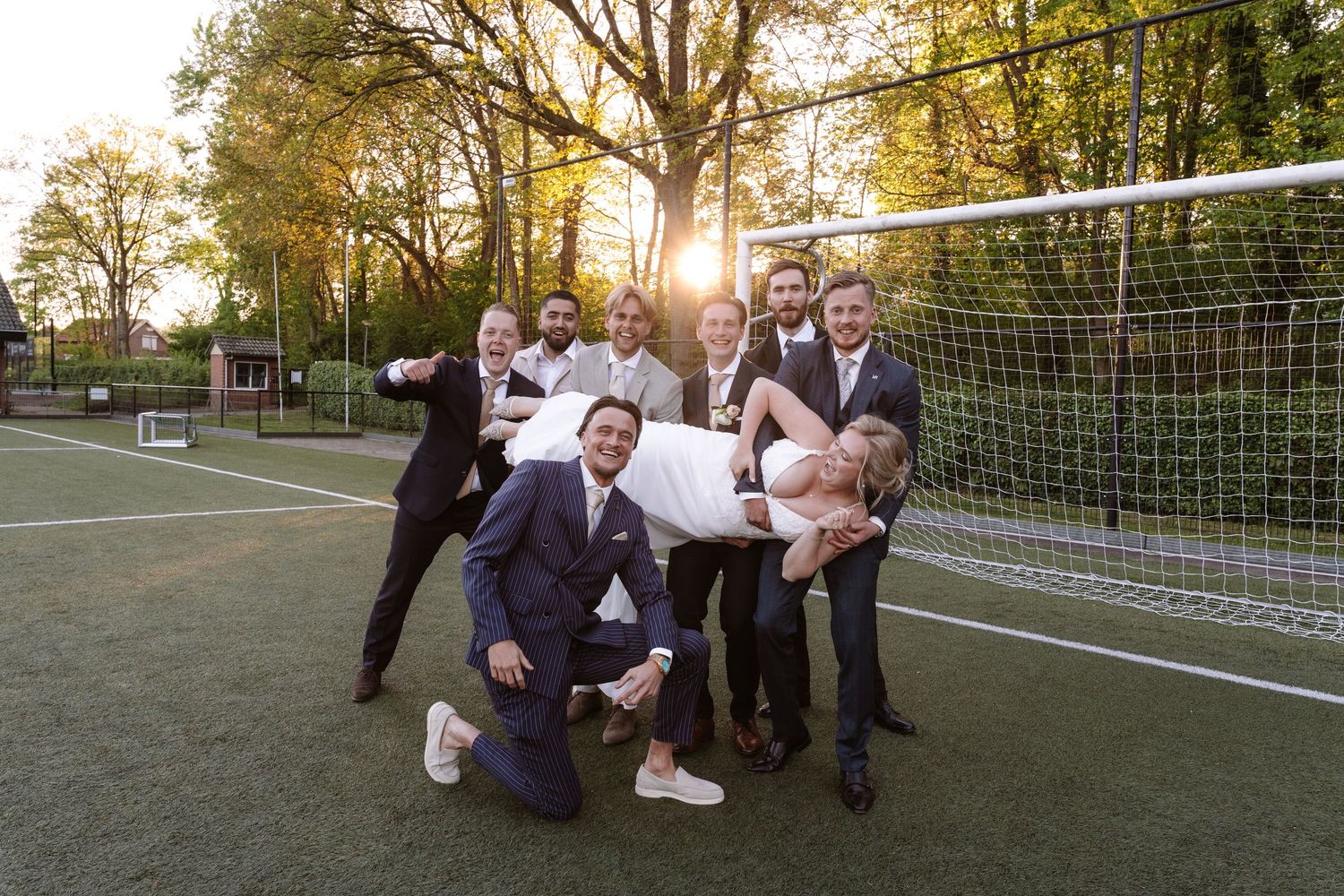 Groomsmen lifting bride on soccer field at golden hour sunset during outdoor wedding photo session.