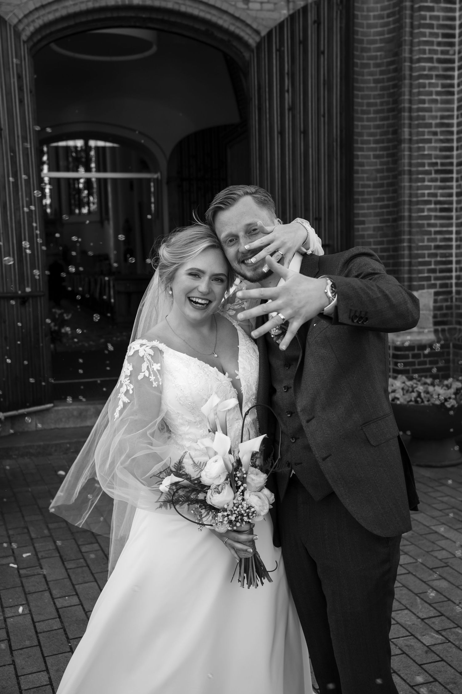 Black and white photo of a smiling bride and groom showing wedding rings outside a brick church.