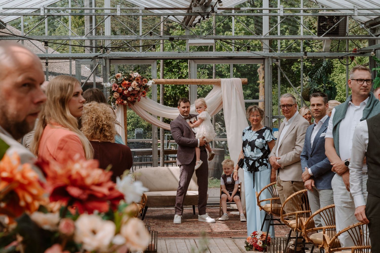 Wedding ceremony in a greenhouse with couple at altar, floral arrangements, and guests watching in elegant attire.