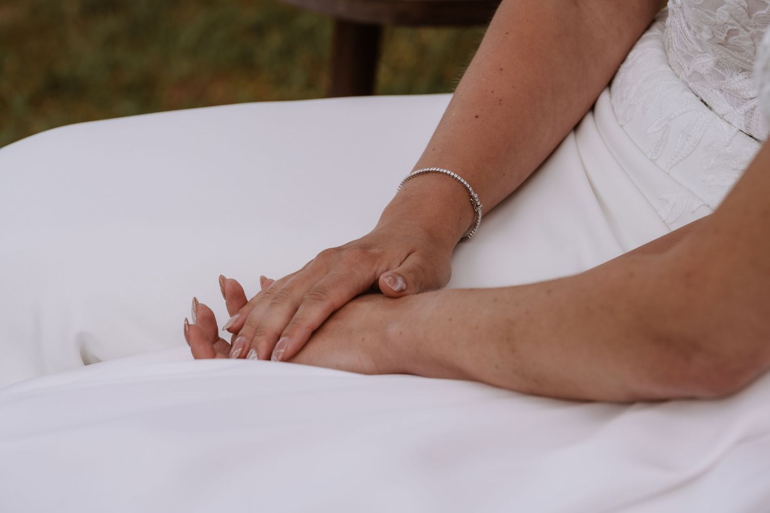Detail shot of hands resting on white wedding dress fabric with a delicate bracelet.