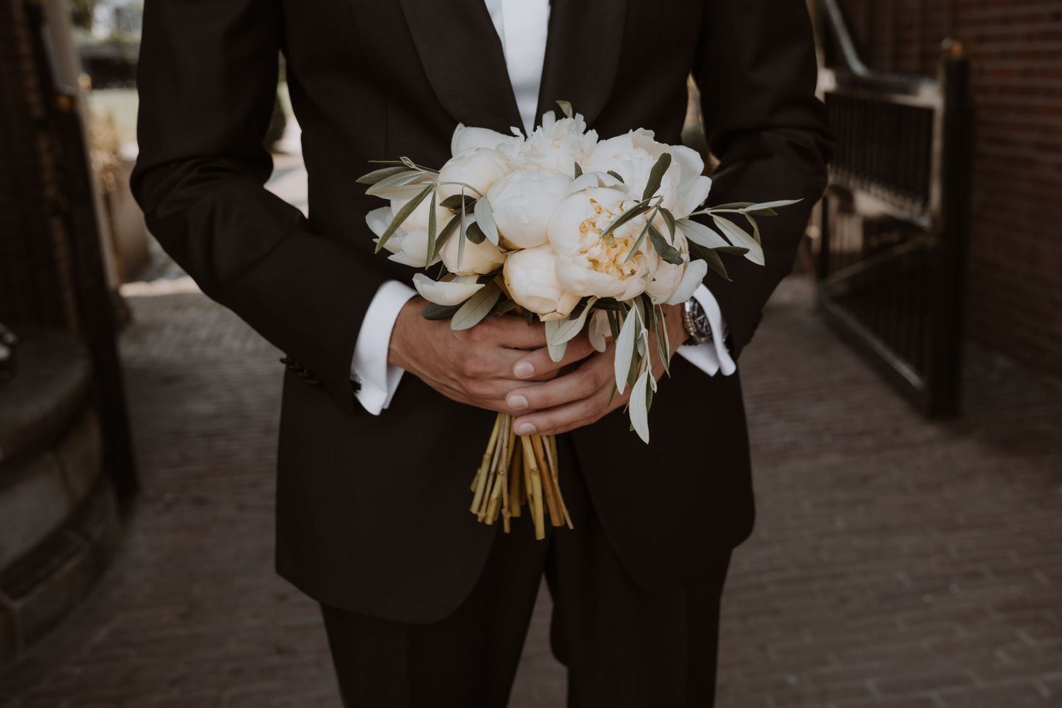 Wedding party members in formal attire holding elegant white floral bouquets against a brick backdrop.