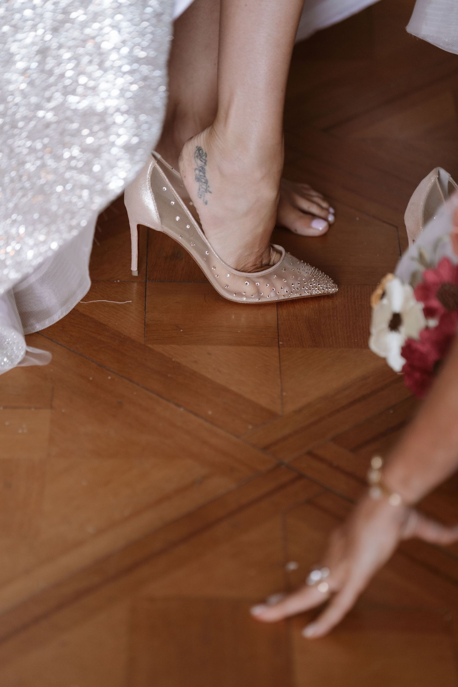 Close-up of sparkly bridal shoes being put on while standing on hardwood floor with pink flowers visible.