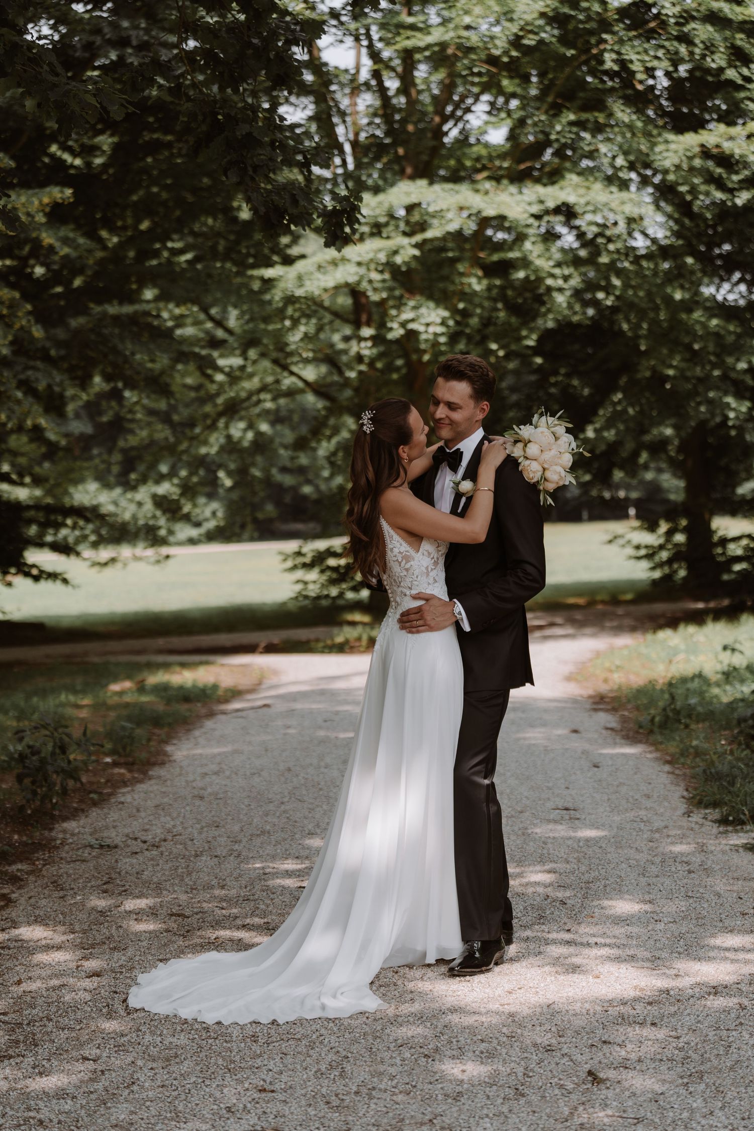 A romantic wedding couple embraces on a tree-lined path in their formal attire.