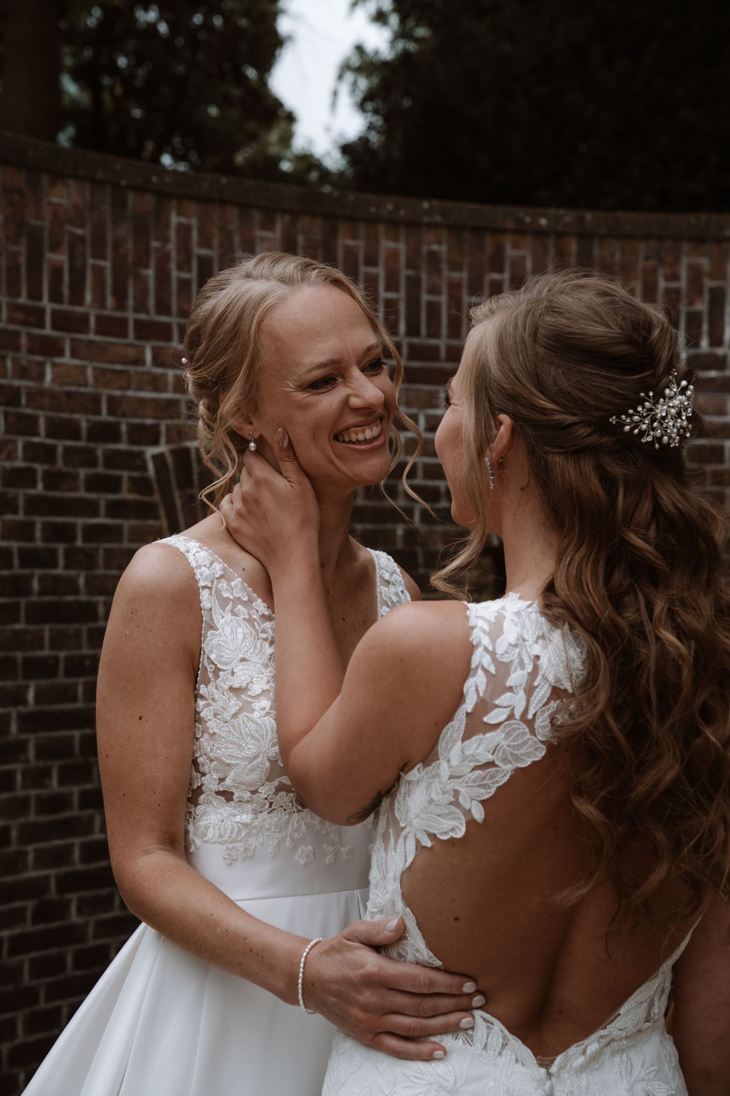 Two people in white wedding dresses share a joyful moment against a brick wall.
