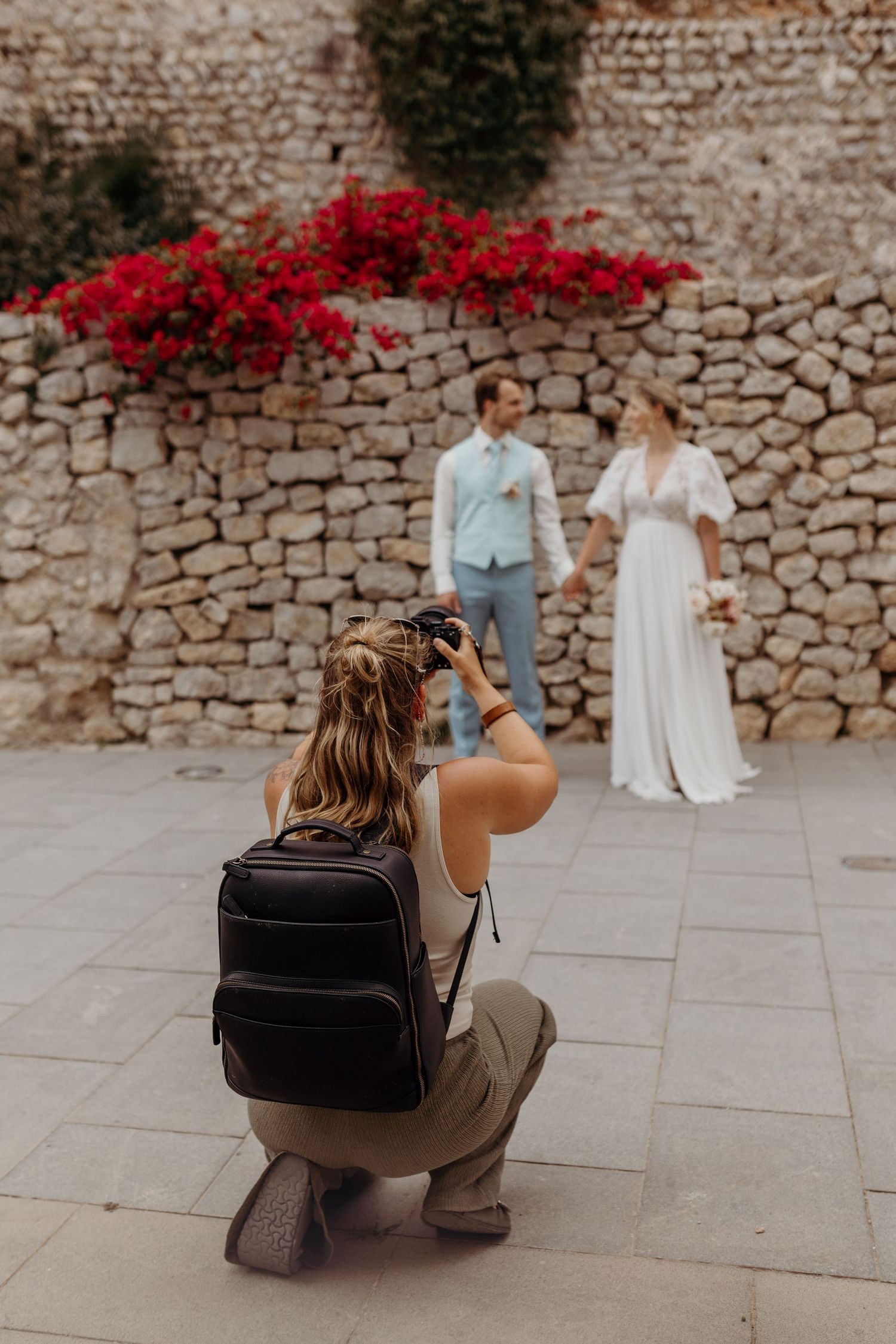 Photographer crouches to capture wedding photos against stone wall with red flowers.