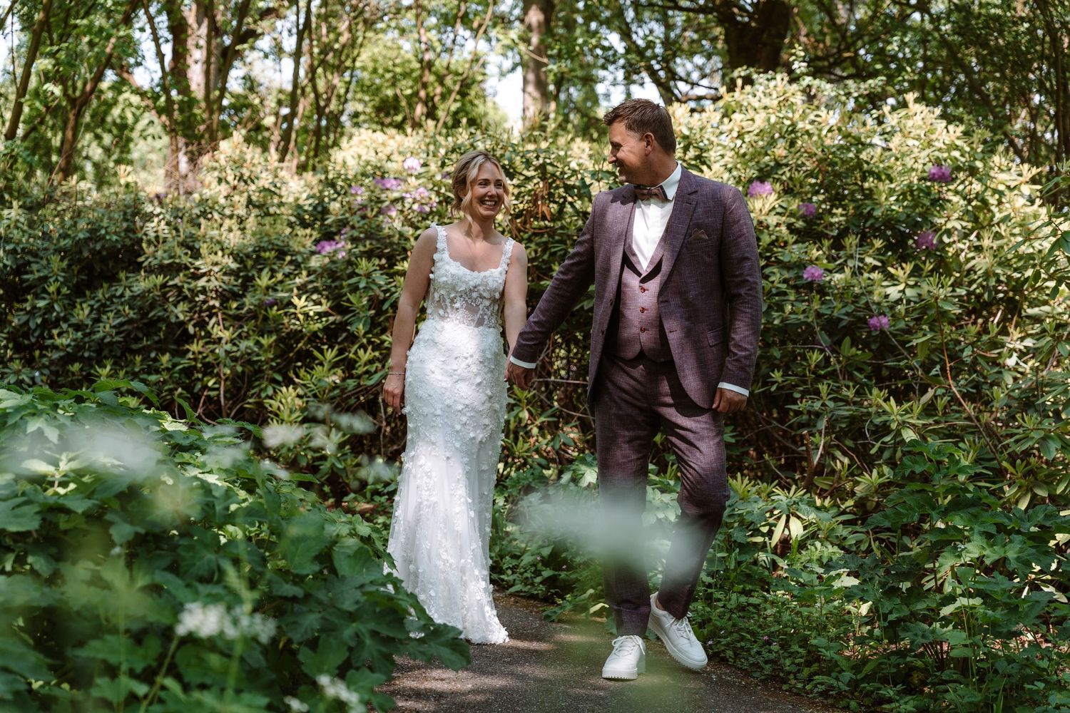 Bride and groom walk hand in hand through a lush green garden with blooming flowers on their wedding day.