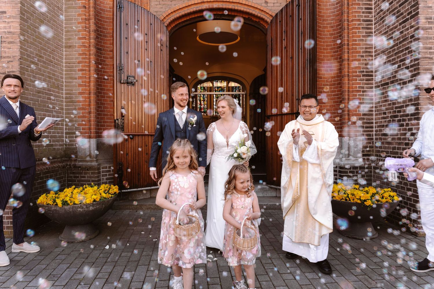 Newlywed couple exits brick church with flower girls, priest, and soap bubbles at joyful outdoor wedding ceremony.
