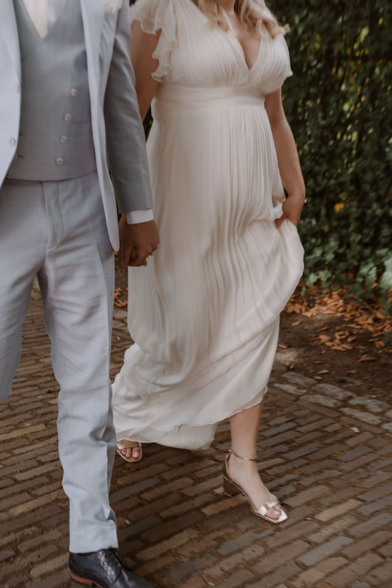 Close-up details of a light gray suit and flowing white wedding dress on a brick pathway.