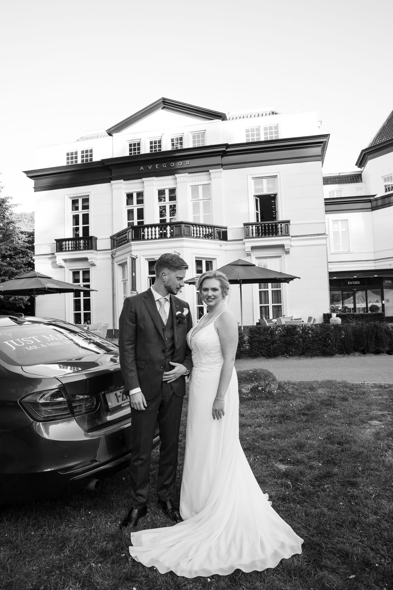 Black and white photo of bride and groom posing outside an elegant manor house beside a Just Married car.