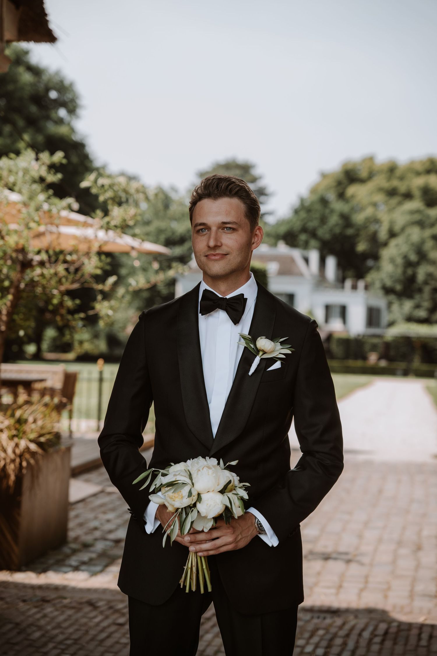 Groomsmen in classic black tuxedos and bowties holding white bouquets outdoors.