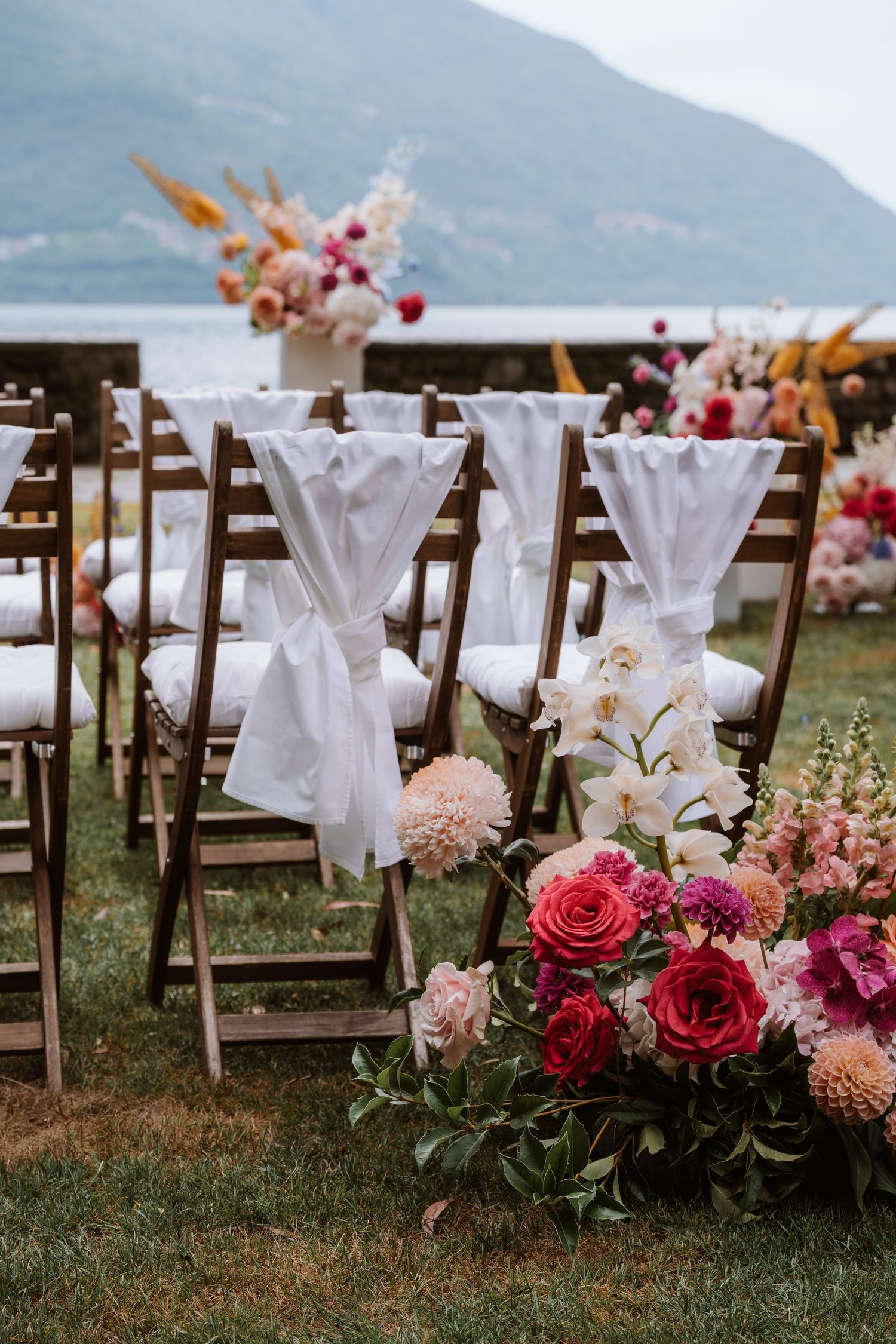Wooden folding chairs with white fabric bows line up at an outdoor wedding ceremony with pink and red floral arrangements.