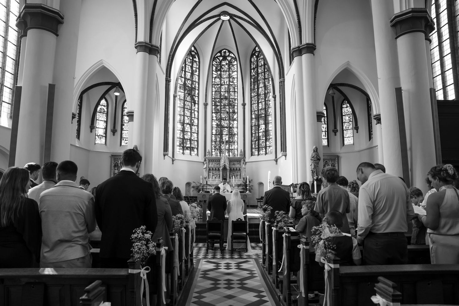 Black and white photo of wedding guests standing in church pews during a ceremony with gothic arched windows and altar.