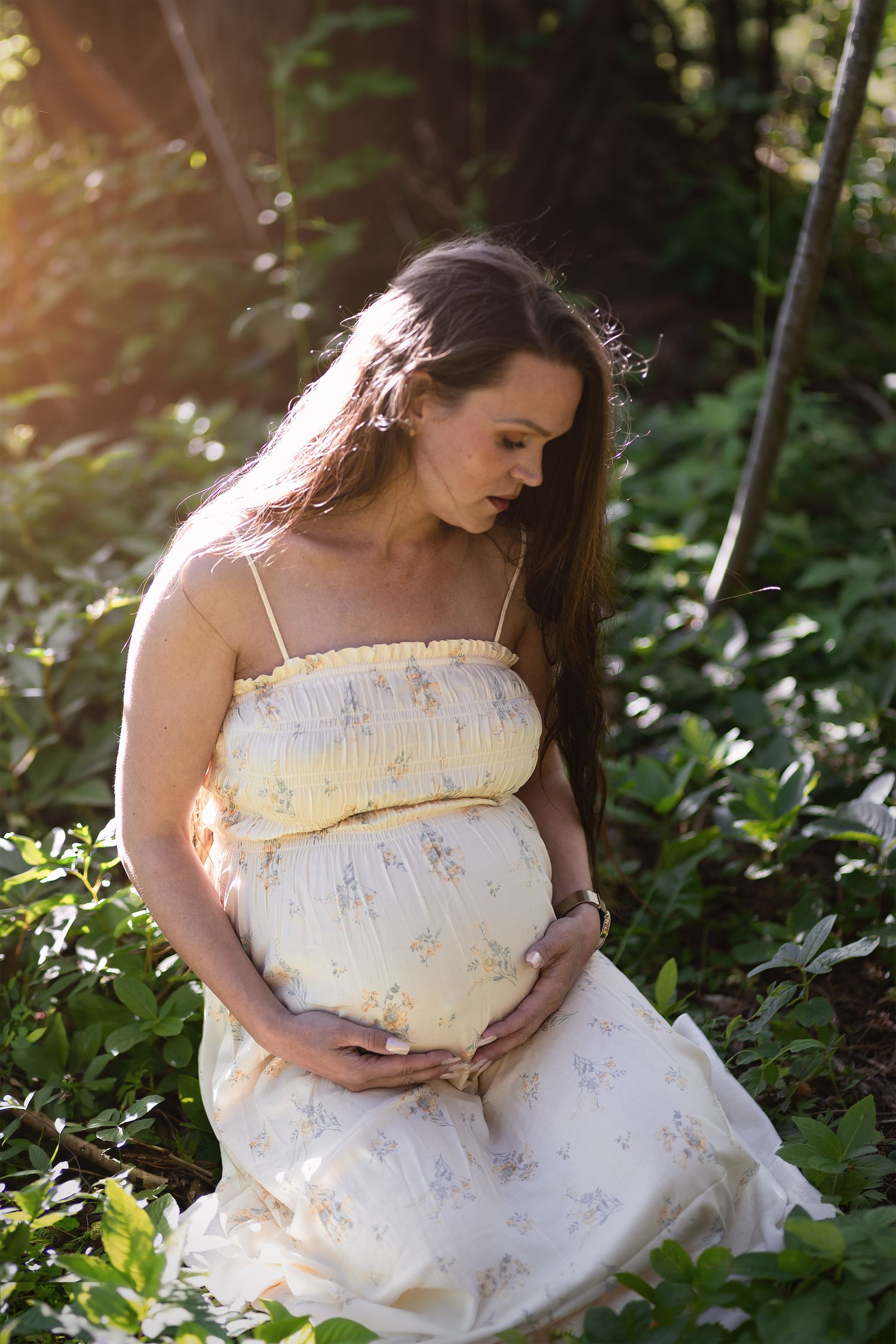 Pregnant woman in yellow floral dress cradling her baby bump while sitting outdoors in lush green garden.