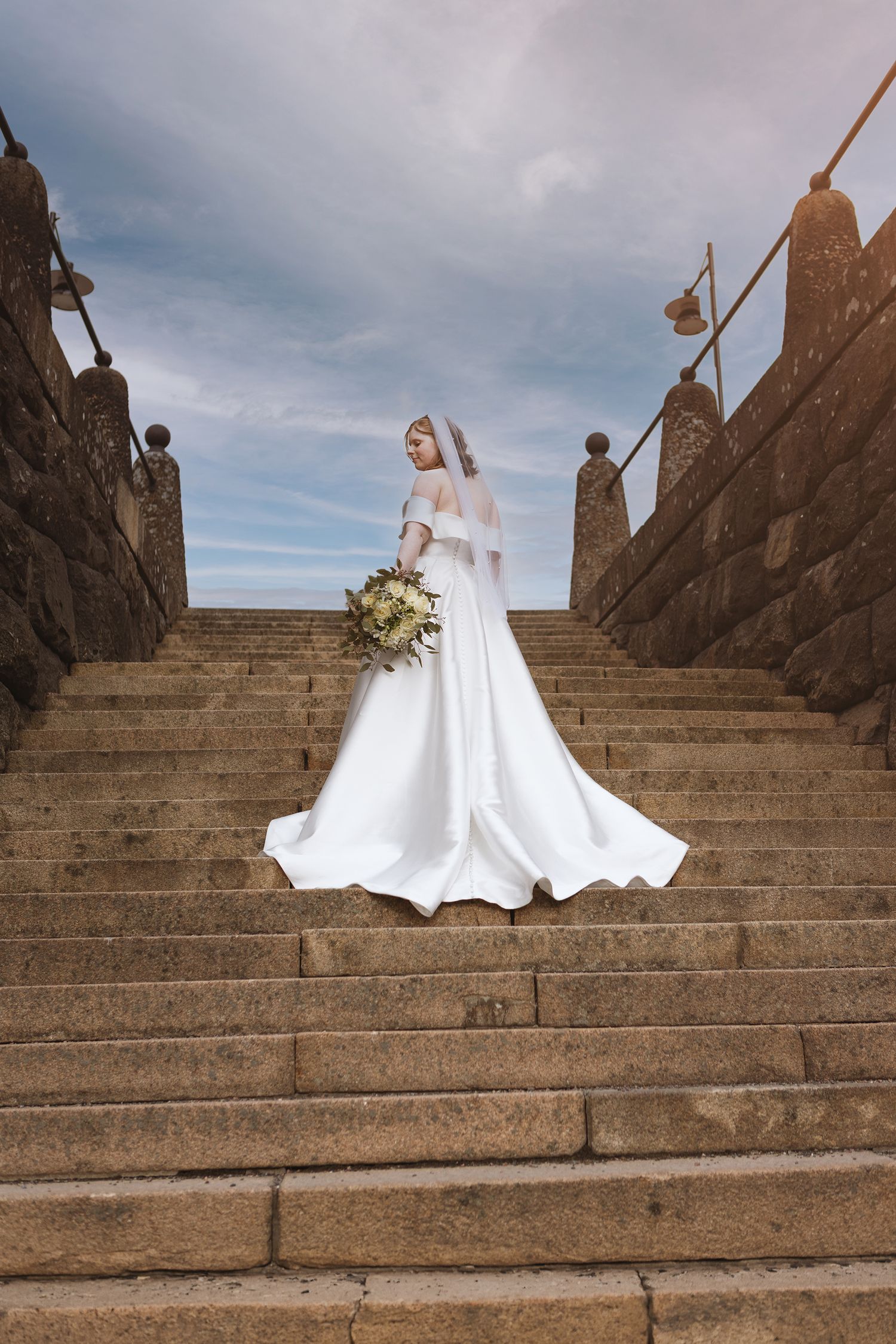 Bride in white gown and veil holding bouquet stands on stone staircase under cloudy sky.