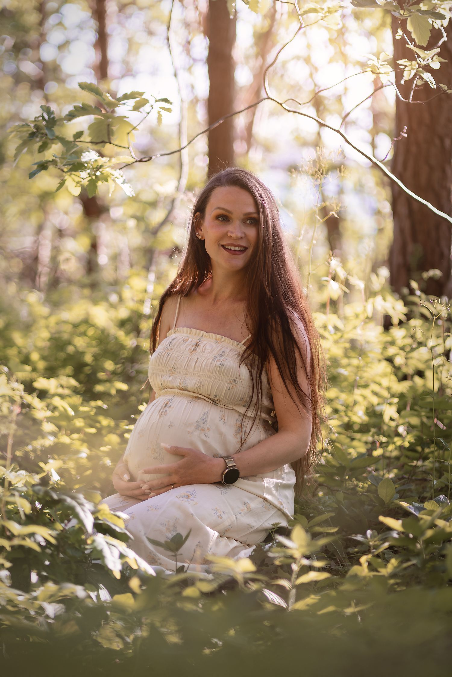 Smiling pregnant woman in white dress sitting among green forest foliage in warm sunlight.