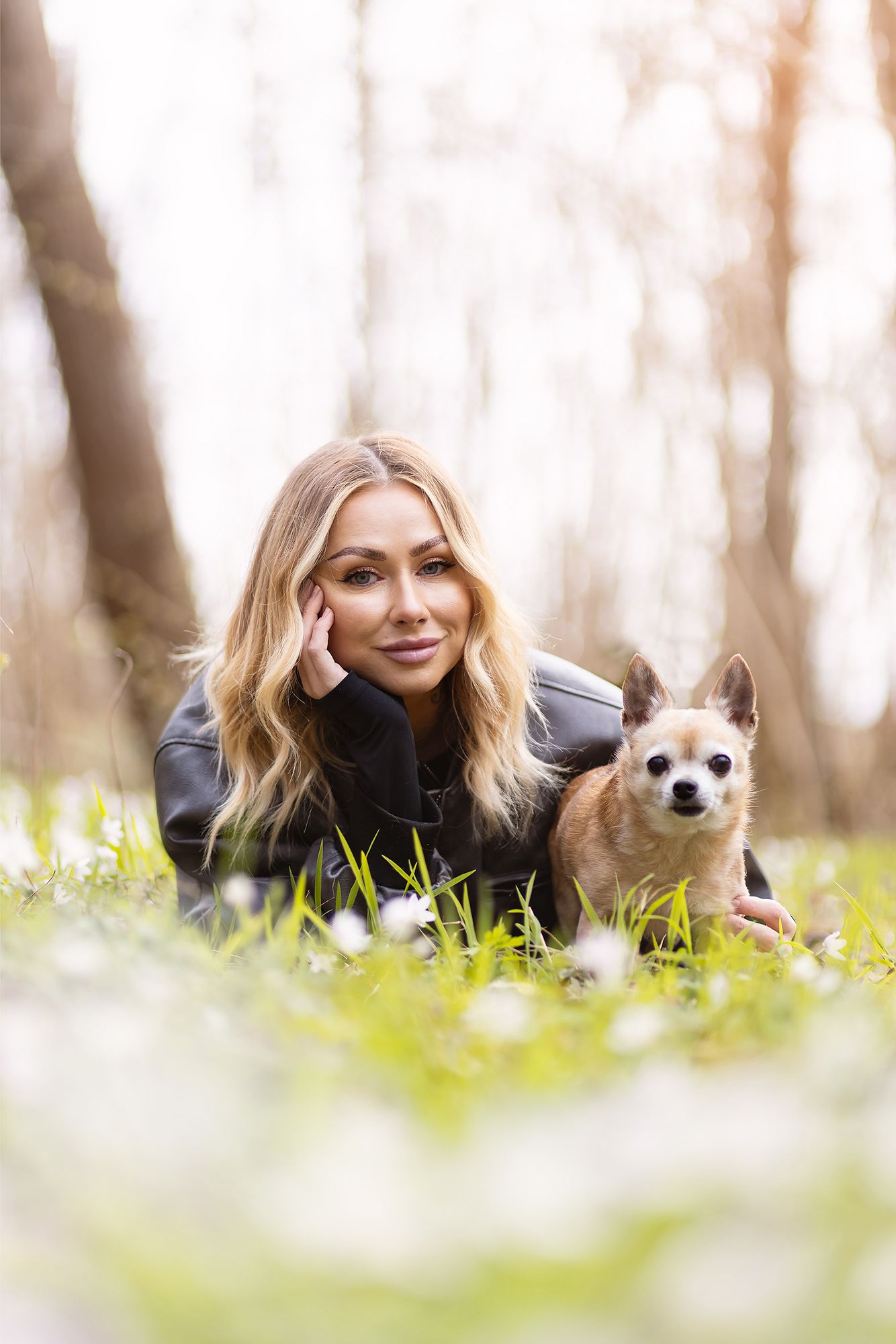 Blonde woman in black leather jacket lying on grass with small Chihuahua dog in a sunlit spring forest.