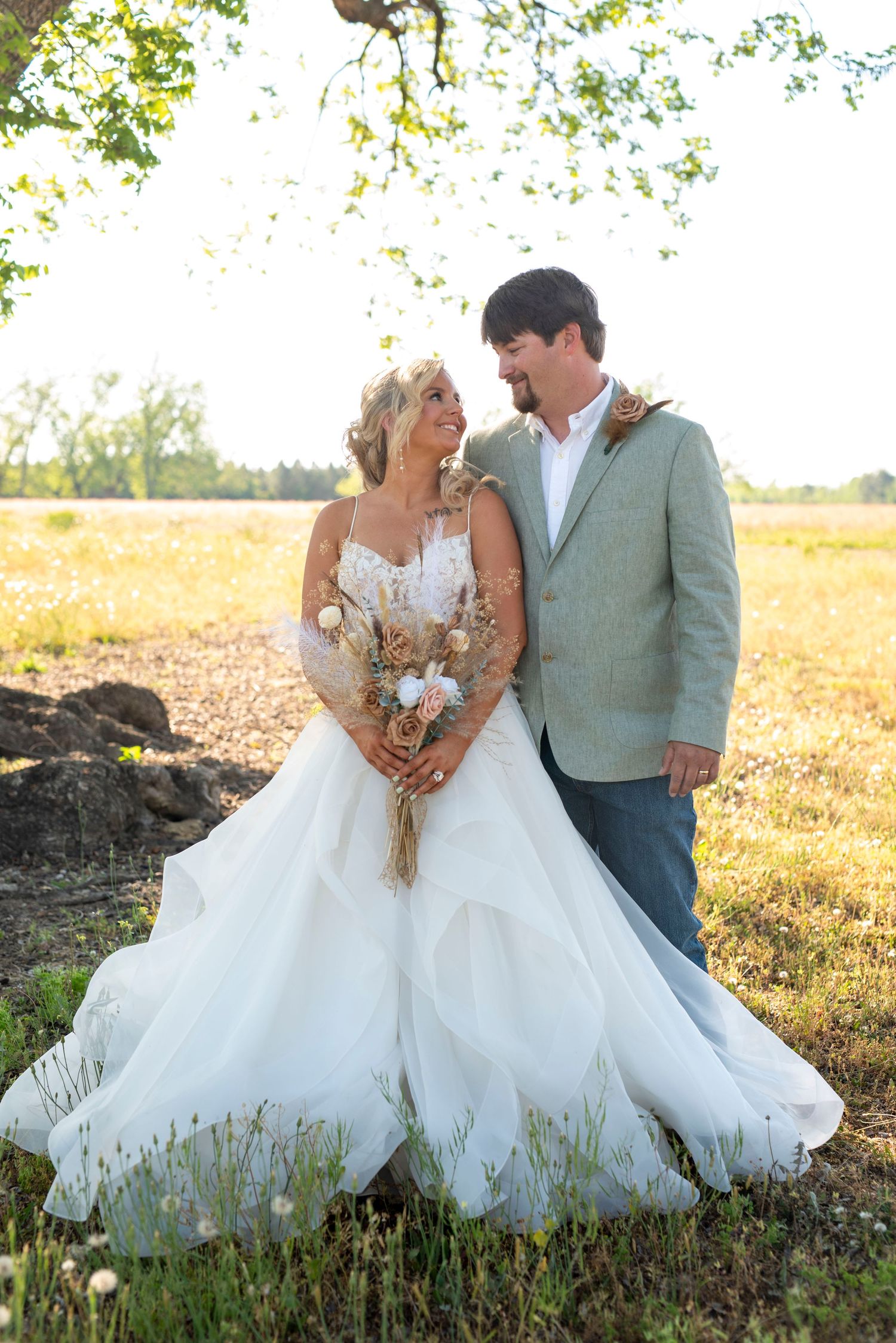 Bride and groom standing together beneath a tree in a sunlit field, surrounded by golden afternoon light.