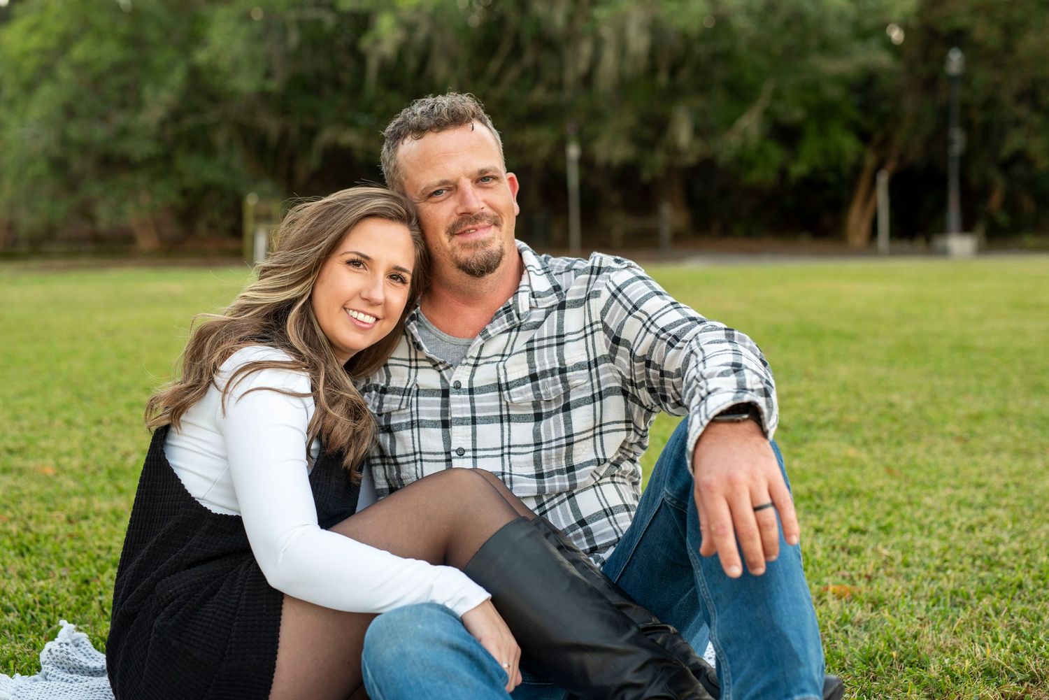 Couple sitting together on the grass during an outdoor portrait session on River Street in Savannah, smiling and leaning into each other.