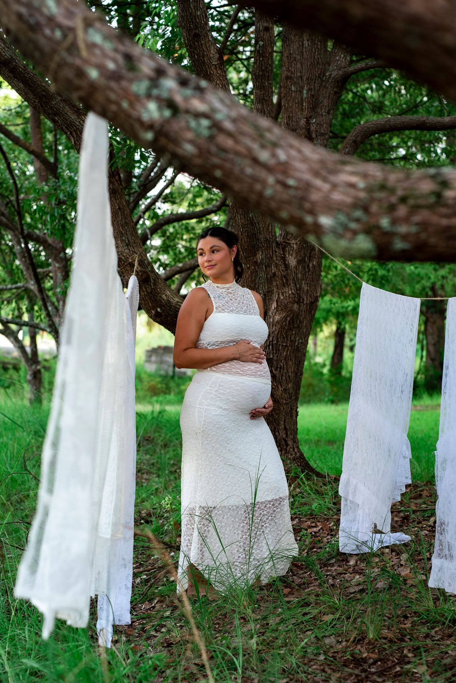 Expectant mother in a white dress cradling her baby bump beside a tree during an outdoor maternity session.