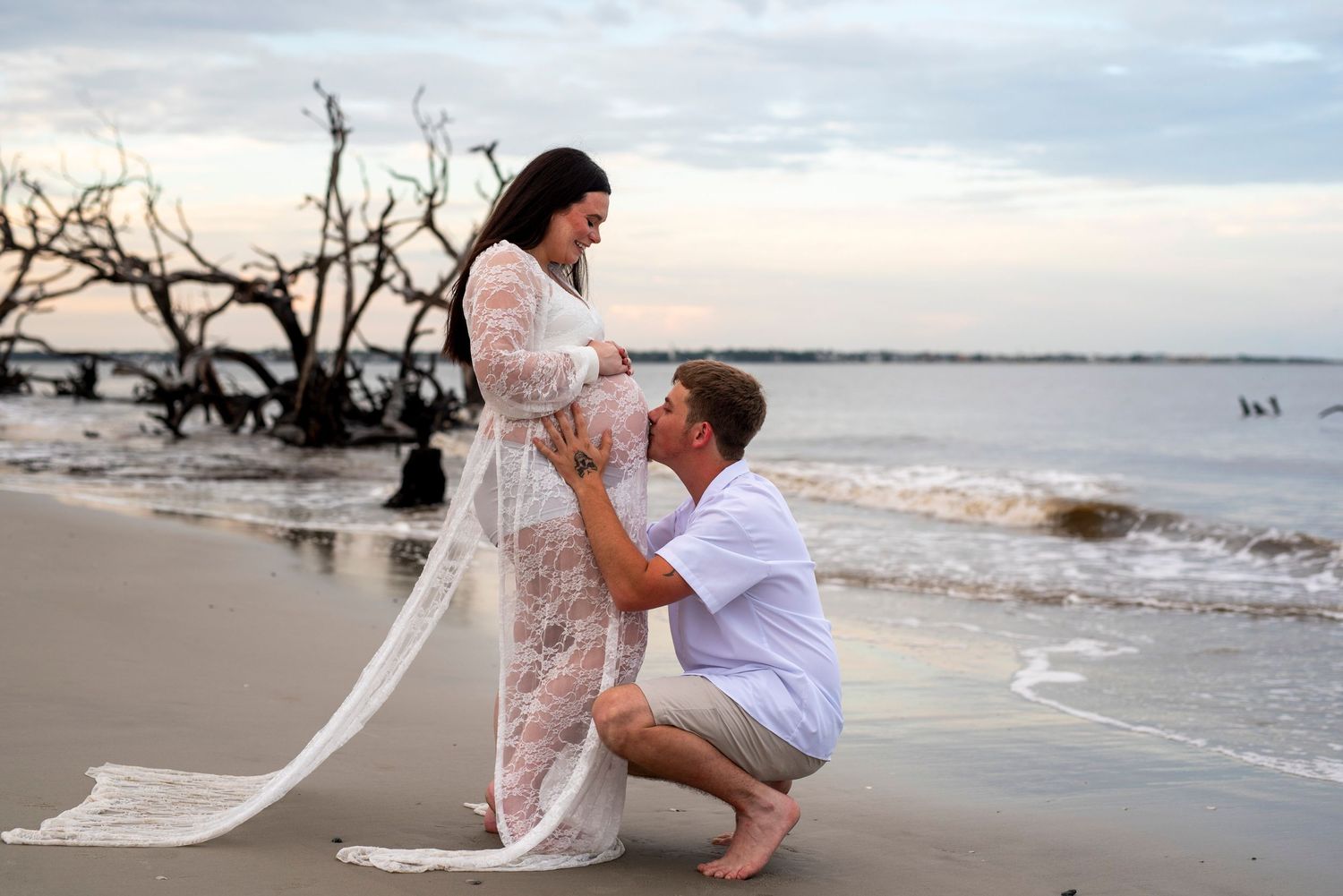 Expectant father kissing his pregnant partner’s baby bump on Driftwood Beach at golden hour during a romantic maternity photography session.