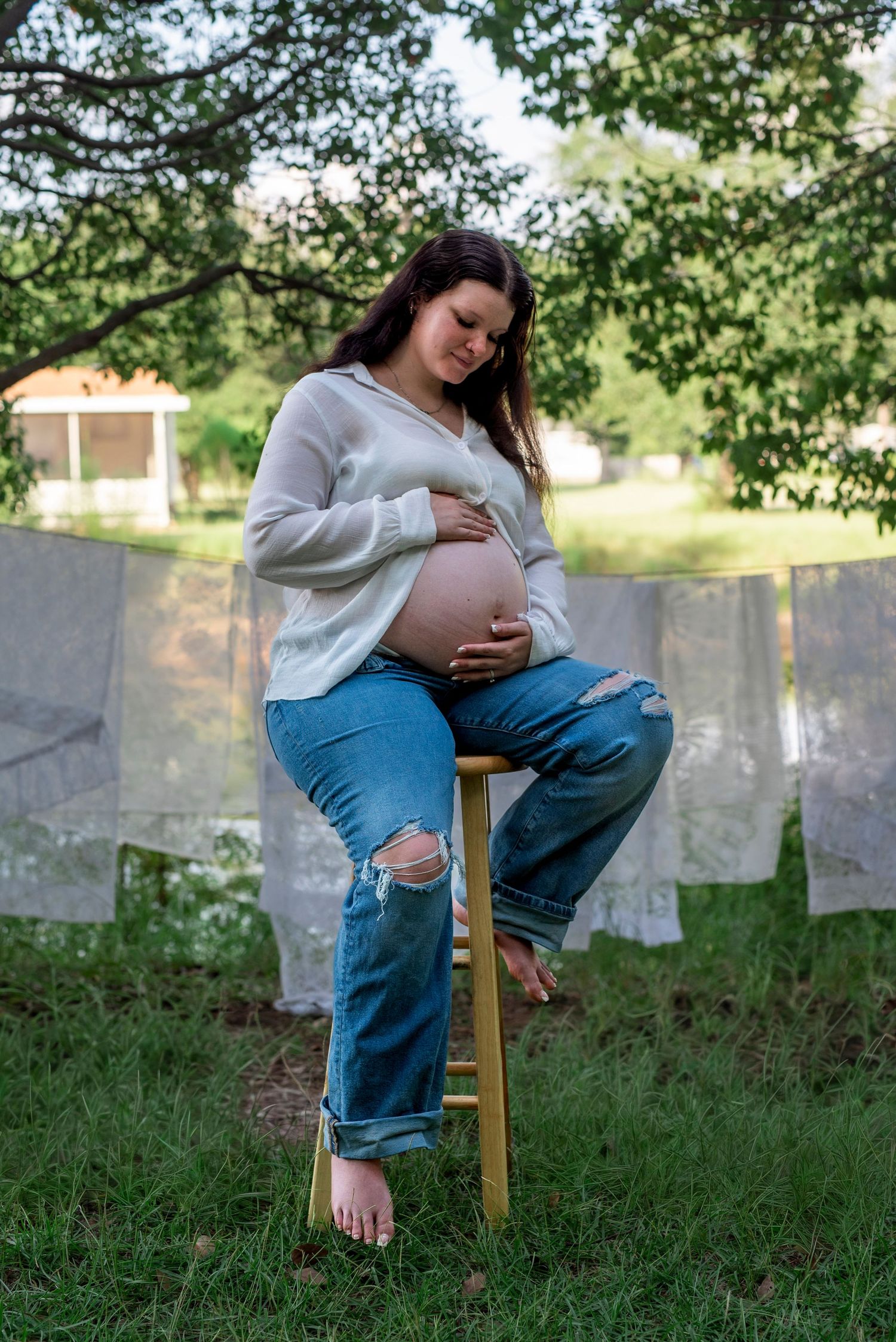 Expectant mother sitting on a stool outdoors with delicate white lace draped behind her during a maternity photography session.
