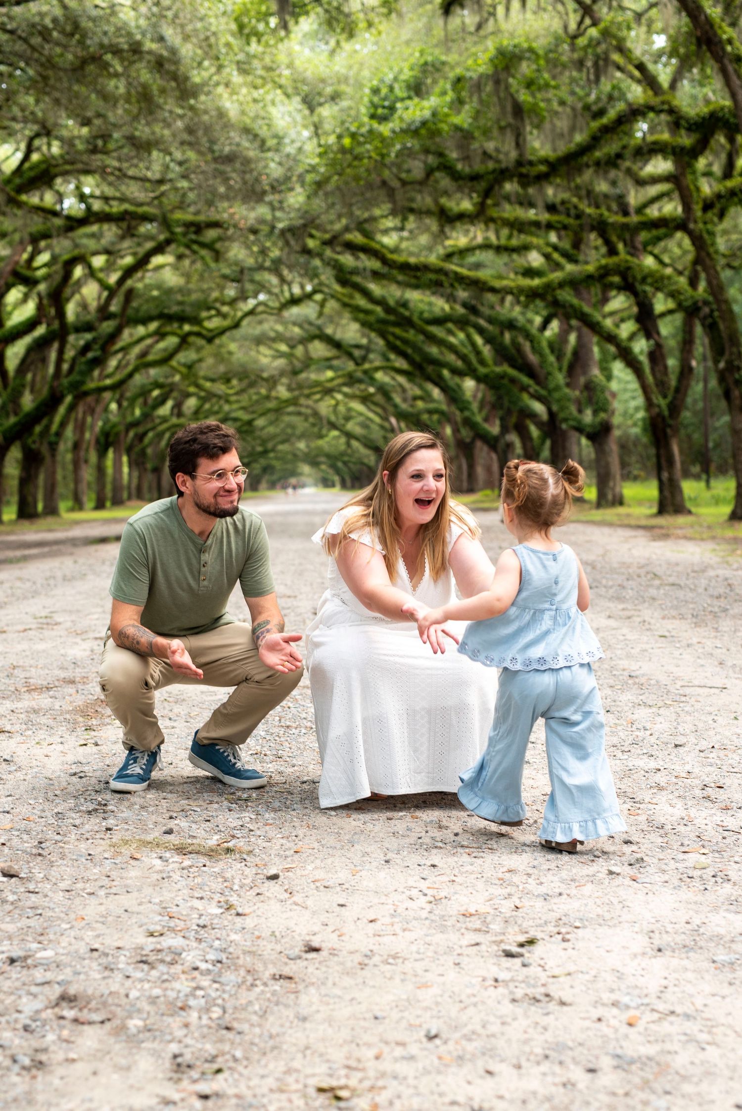 Family portrait of parents laughing and playing with their young daughter on a tree-lined outdoor path at Wormsloe Historic Site in Savannah