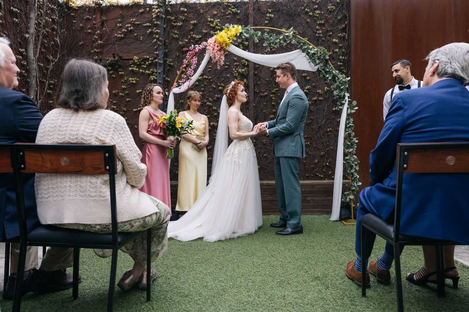 Bride and groom exchange vows under a floral arch at an intimate outdoor wedding ceremony with guests seated nearby.