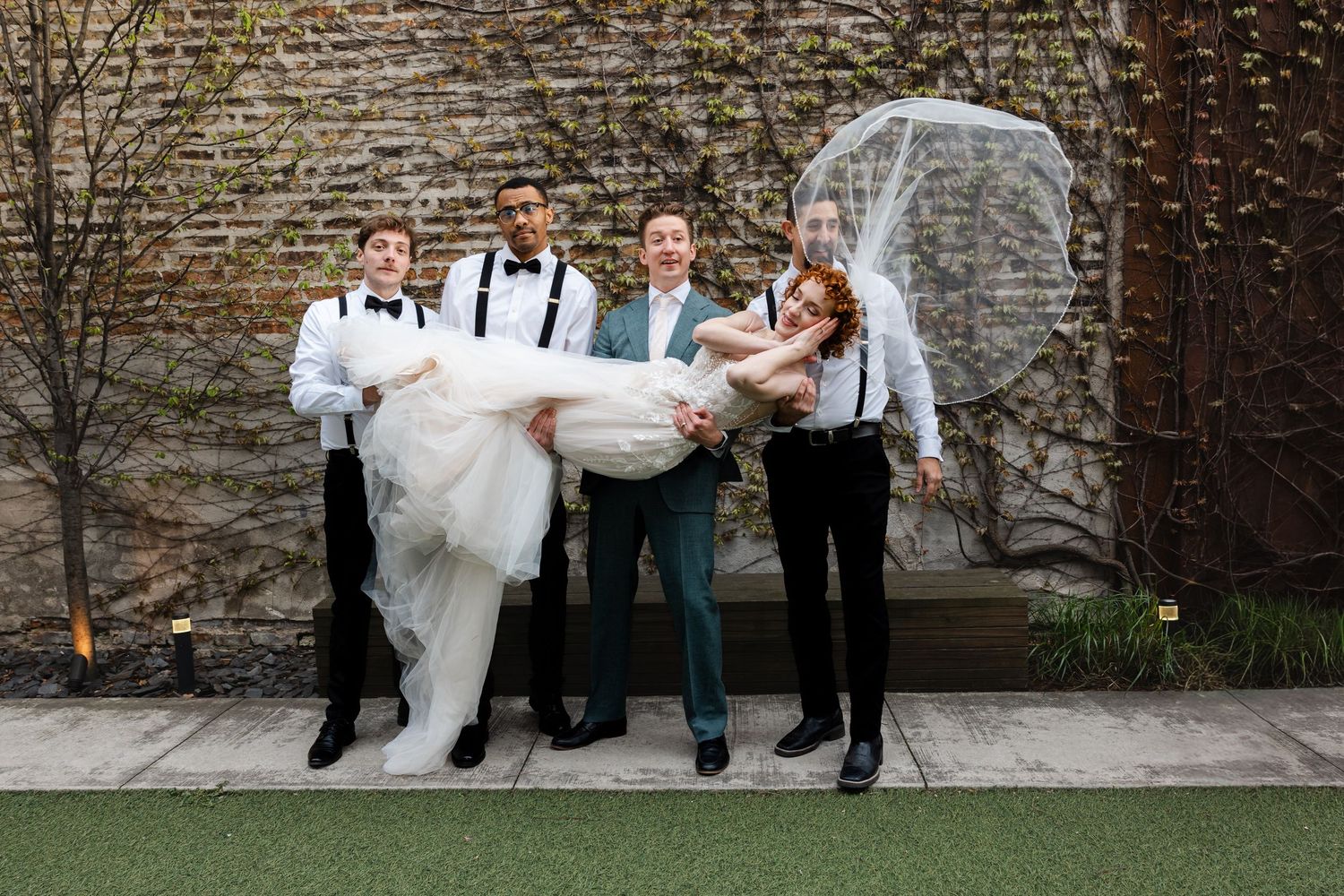 Three groomsmen in suspenders lifting smiling bride in white dress outdoors.