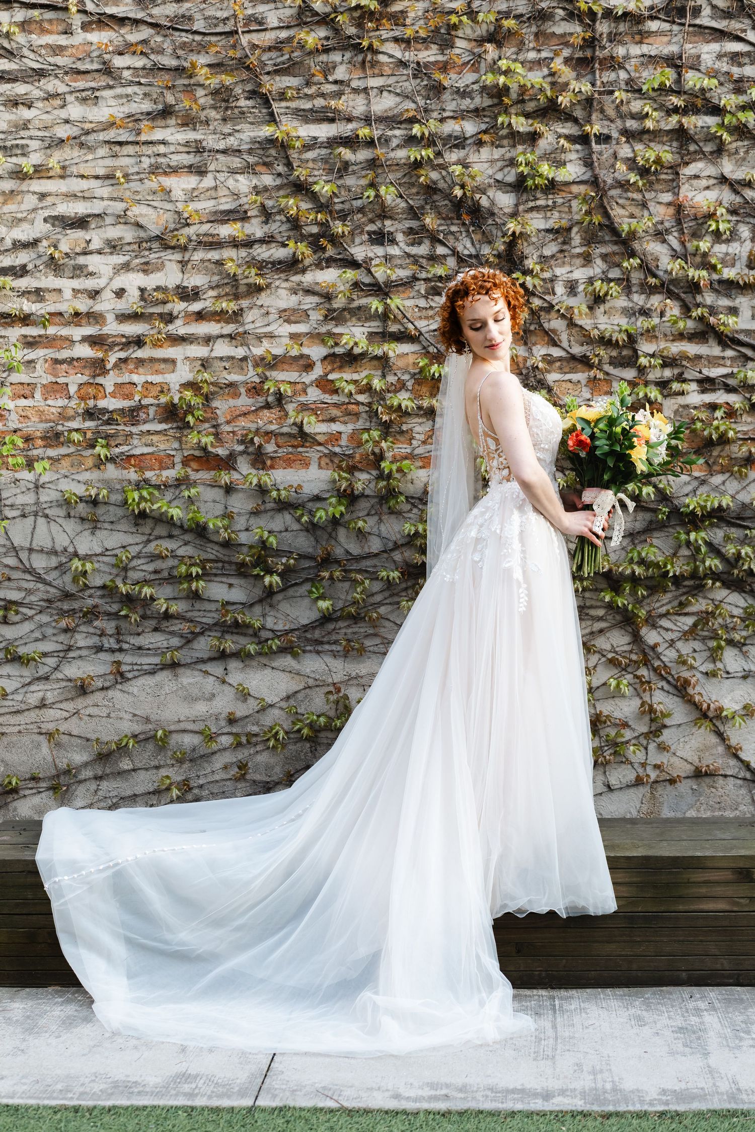 Elegant red-haired bride in flowing white tulle gown with long train holds colorful bouquet before ivy brick wall.