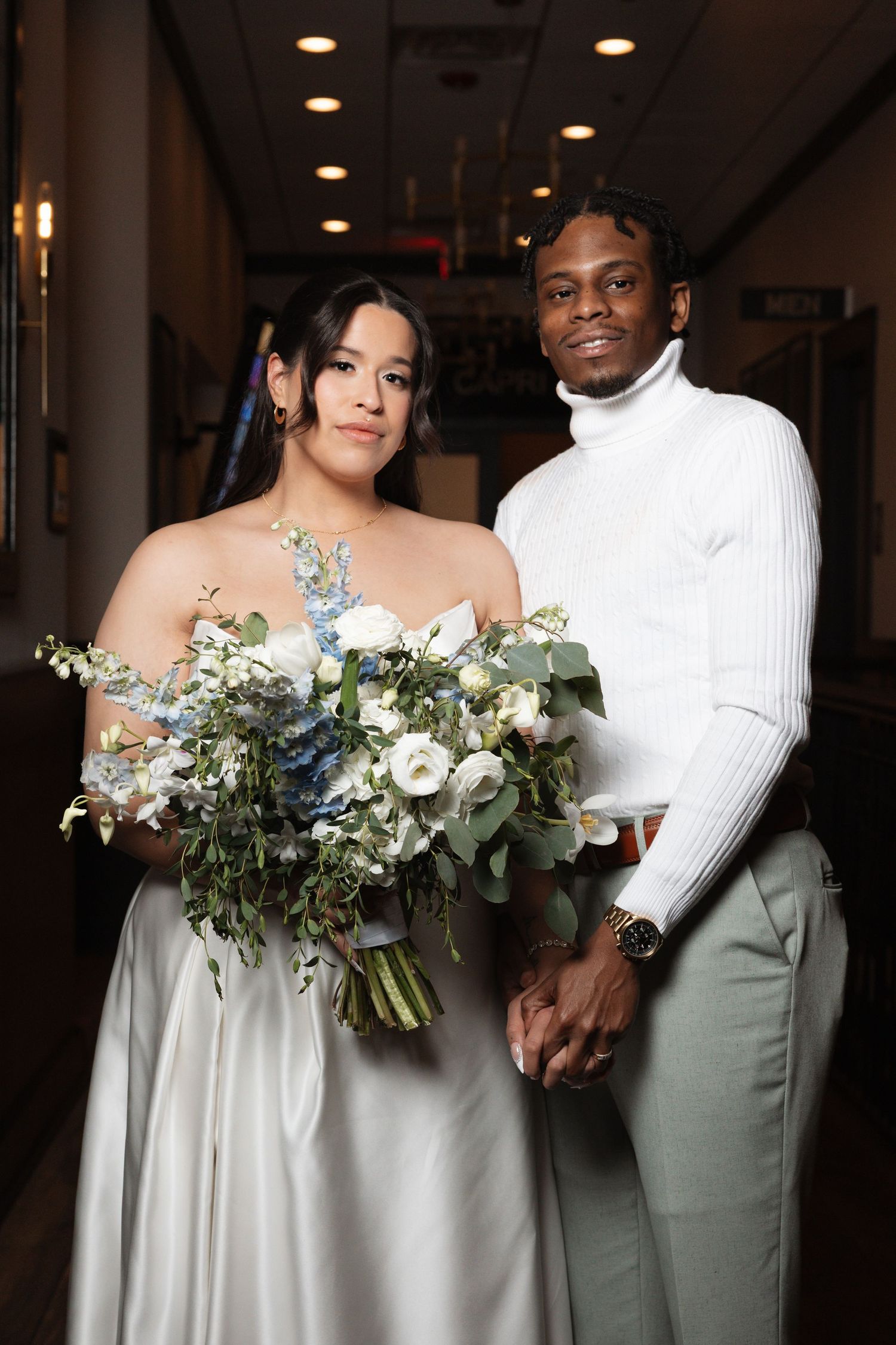 A bride holding a blue and white floral bouquet poses with a groom in a white turtleneck at an indoor wedding venue.