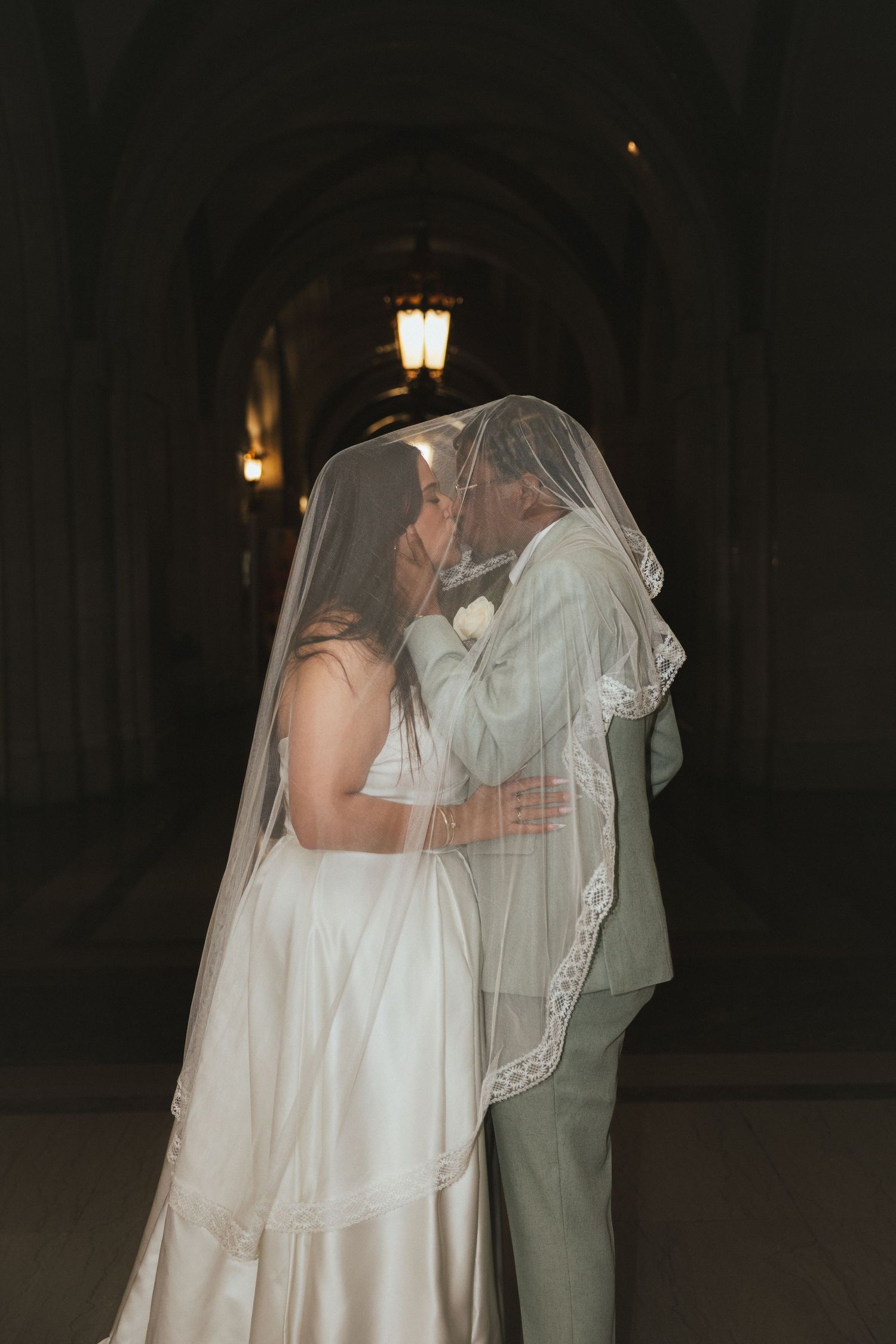 Bride and groom share romantic kiss hidden beneath flowing wedding veil in dimly lit venue.