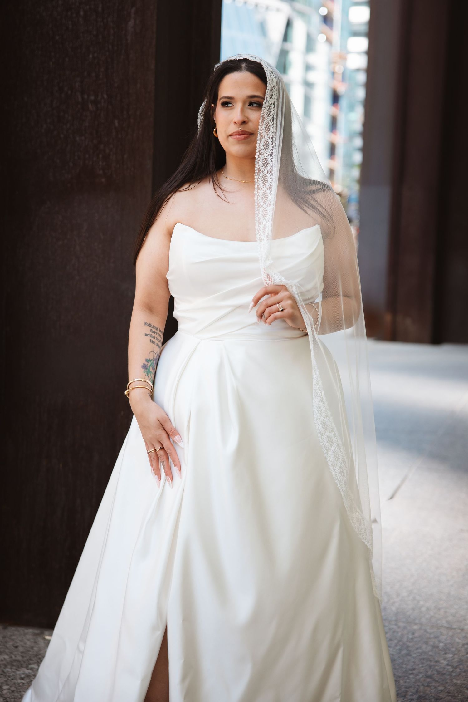 Bride in strapless white satin gown with lace veil and arm tattoo posing between dark columns.