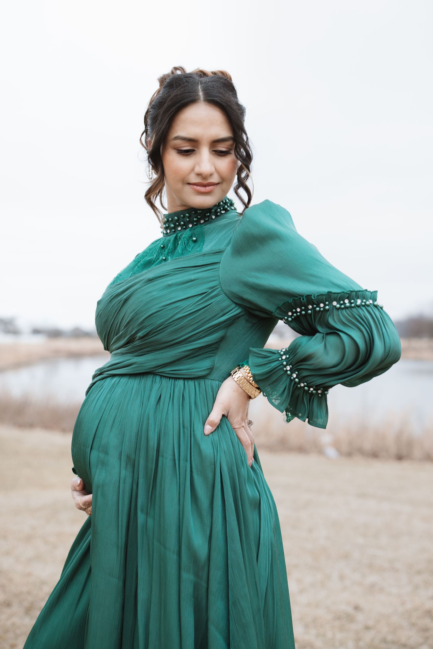 Pregnant woman in emerald green dress with pearl details looking down at baby bump.