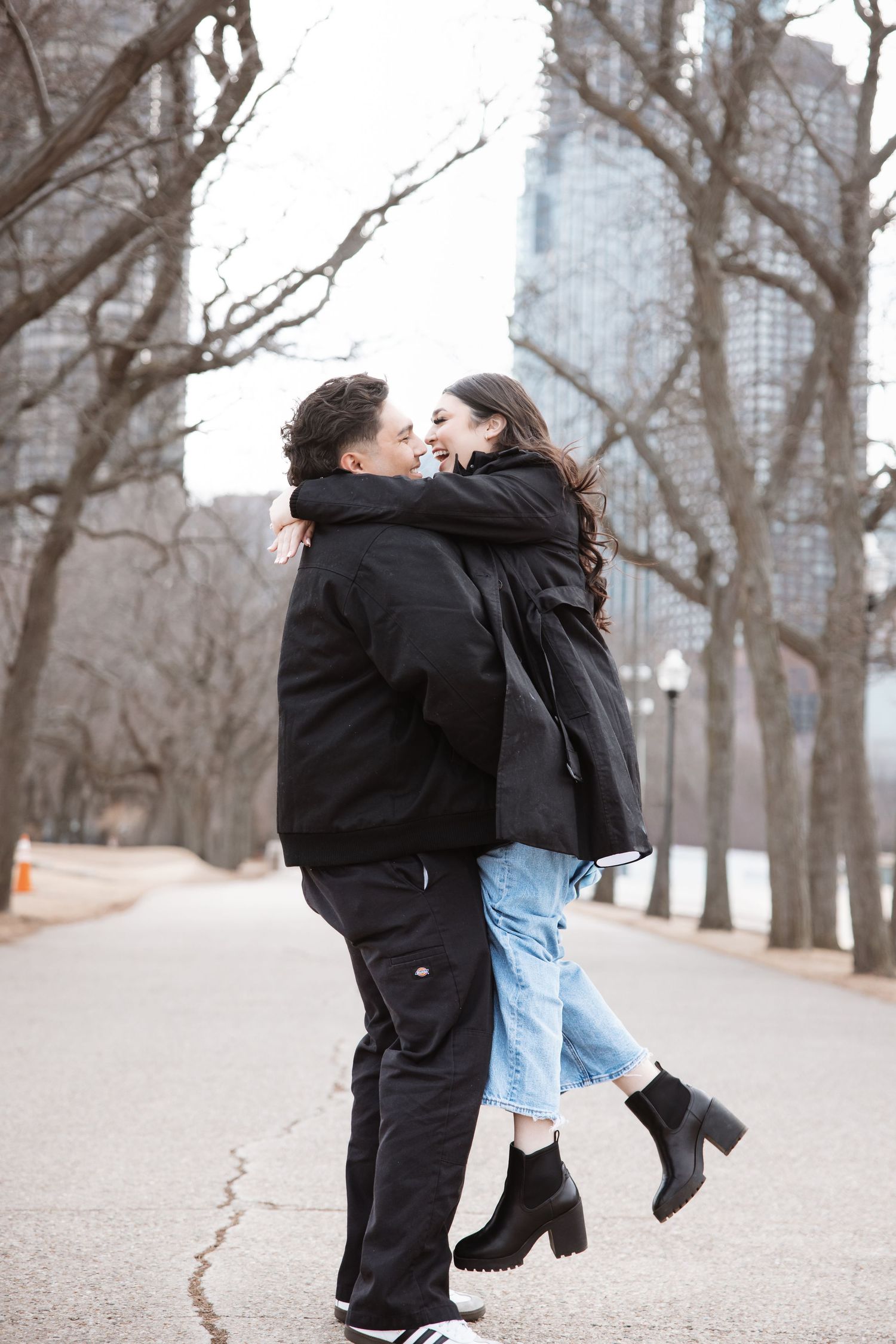 Happy couple embraces and laughs on tree-lined path in winter urban park setting.