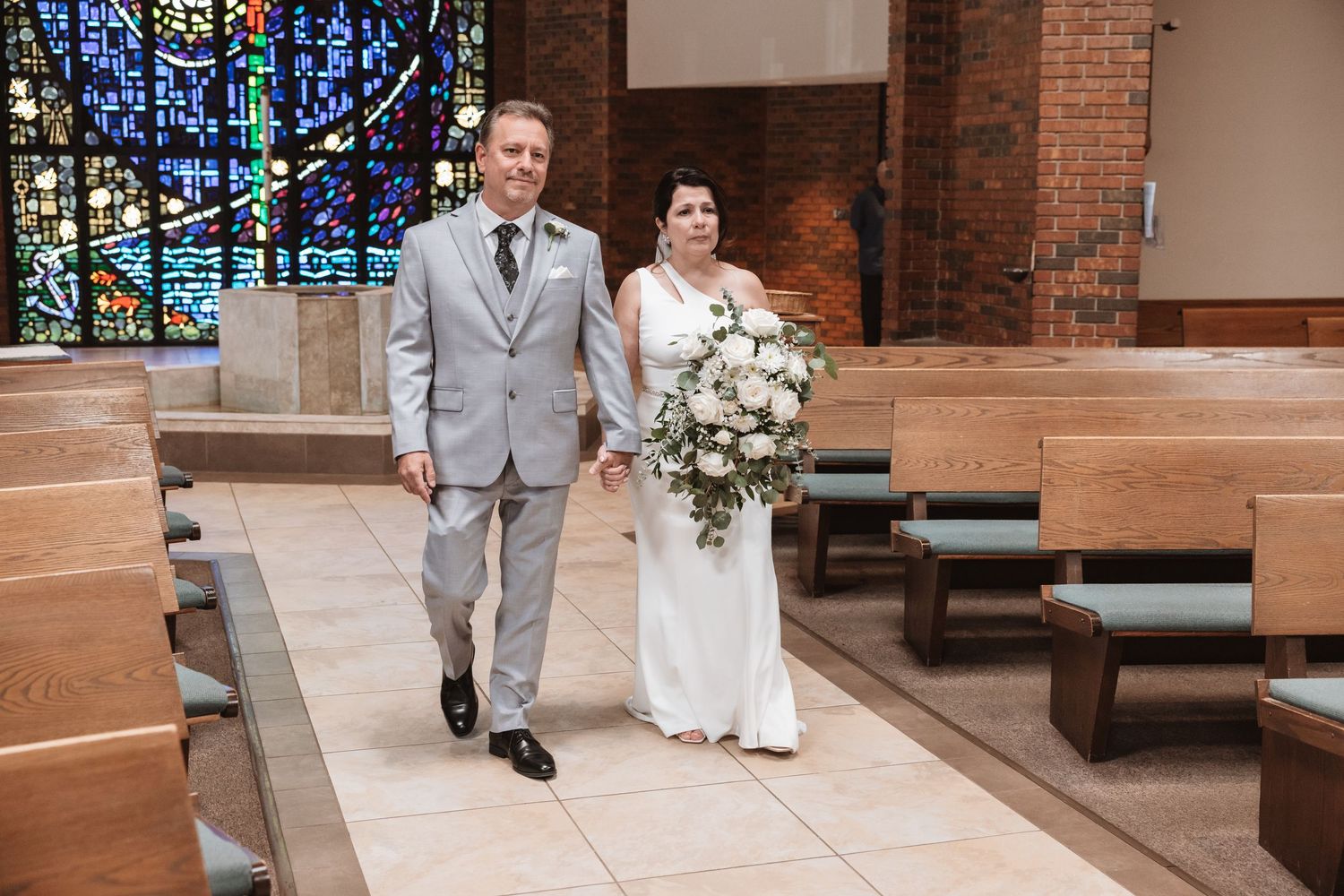 Bride and groom walk hand-in-hand down church aisle, bride holding white floral bouquet, groom in gray suit.