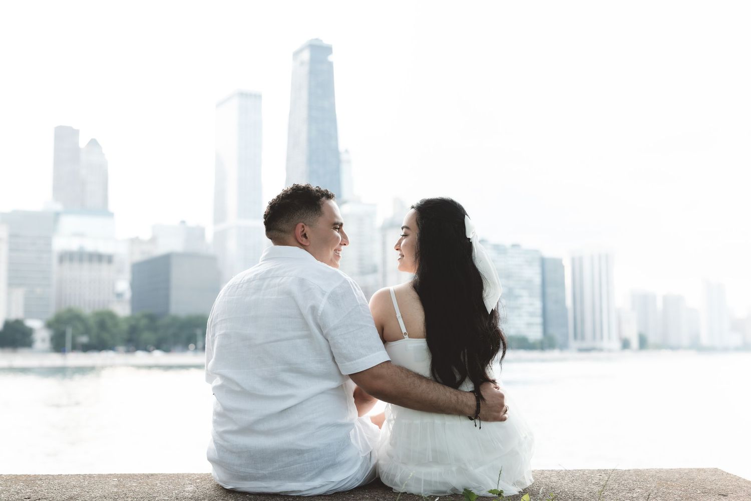Couple sitting together on a waterfront ledge in white outfits, with a city skyline in the background.