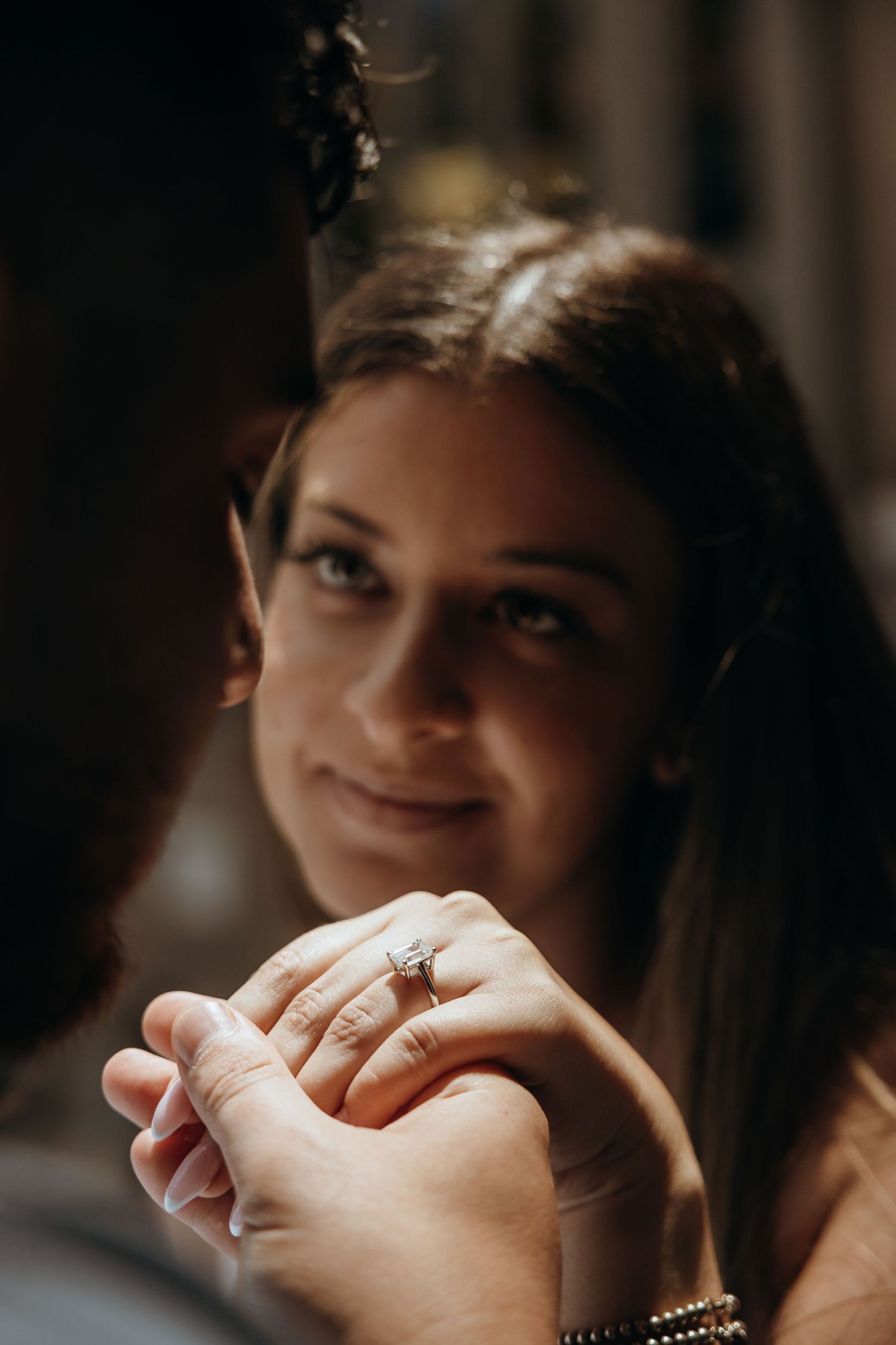 Close-up of smiling bride showing diamond engagement ring while holding hands with her partner in warm light.