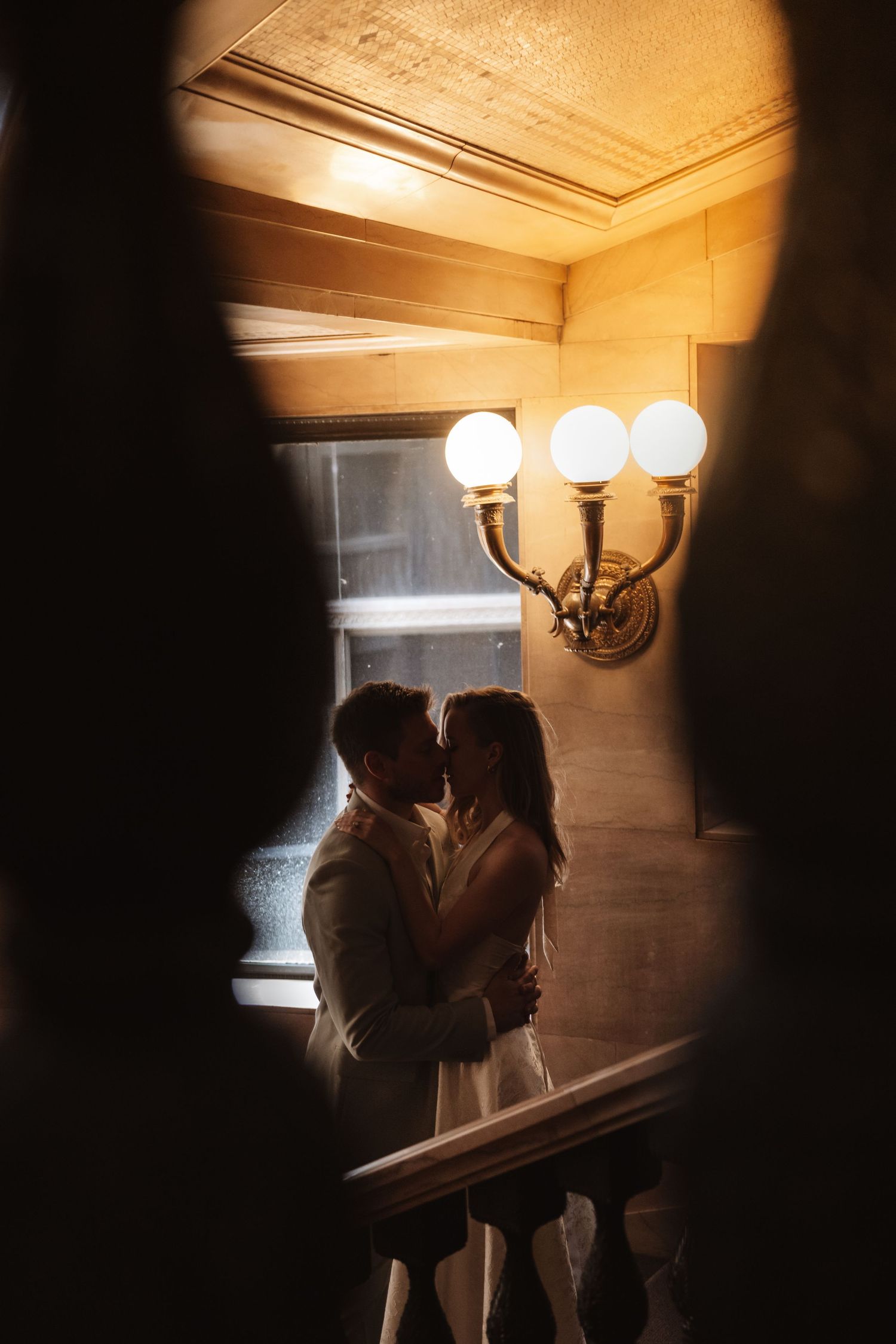 Couple sharing a romantic kiss on a staircase landing, framed by warm wall sconces in an elegant venue.