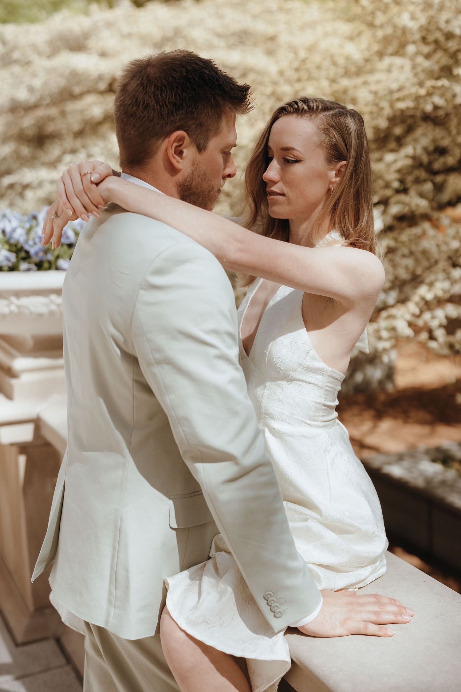 Bride in white mini dress and groom in beige suit share a tender embrace in a blooming garden.