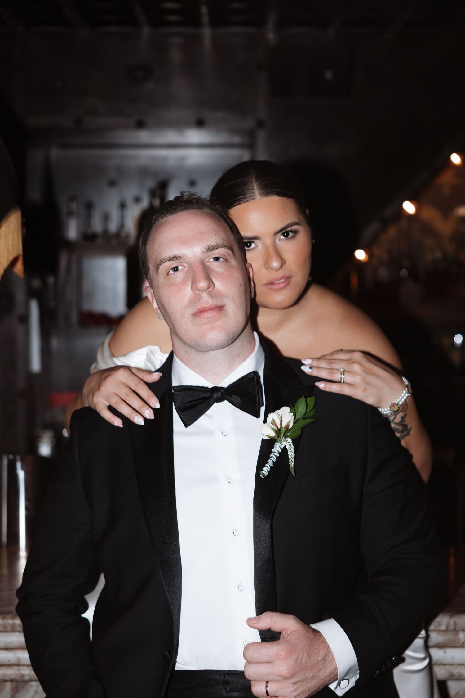 Elegant couple in formal wedding attire, groom in black tuxedo with boutonniere, bride in white gown embracing him from behind.