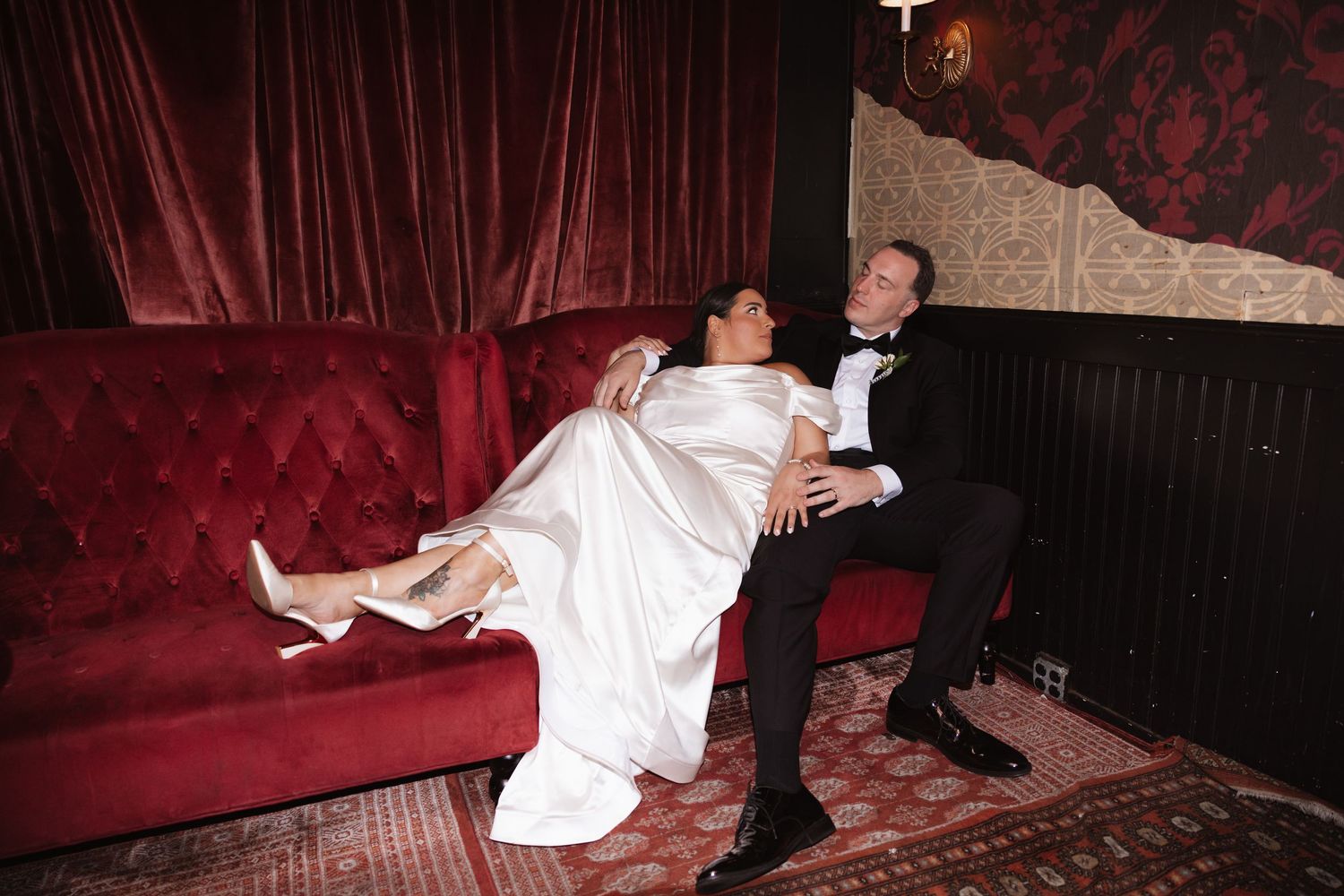 A bride in a white gown is cradled by her groom on a red tufted velvet sofa at their wedding reception.