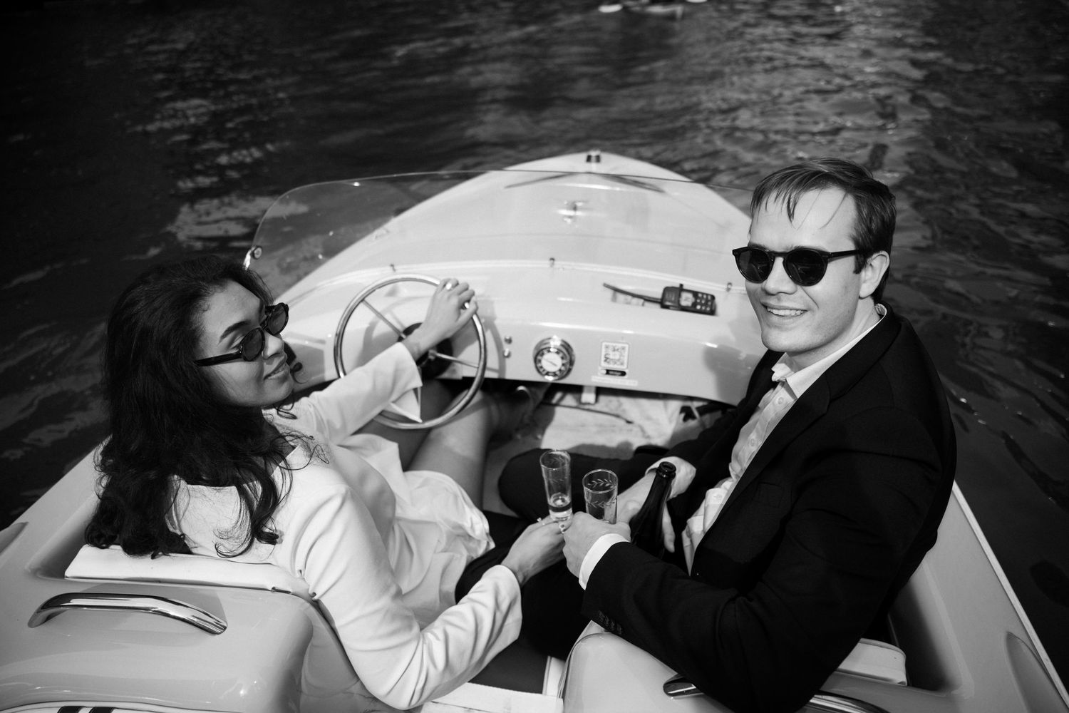 Black and white photo of a smiling couple in sunglasses riding a boat on the water, dressed in wedding attire.