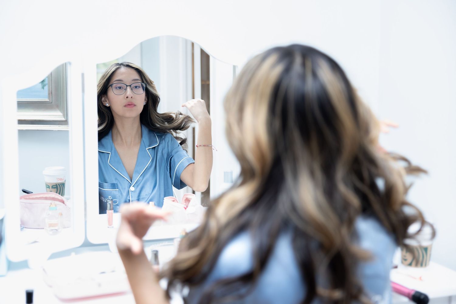 A young woman with highlighted hair examines her reflection in a lighted vanity mirror during wedding prep.