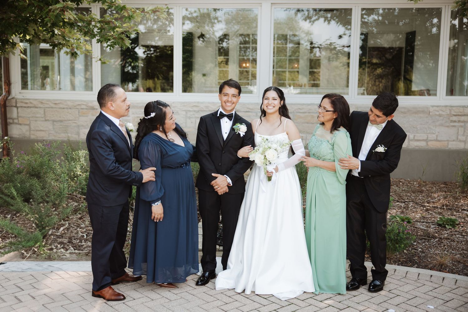 Bride and groom laugh with parents in navy and sage green formal wear at outdoor wedding.