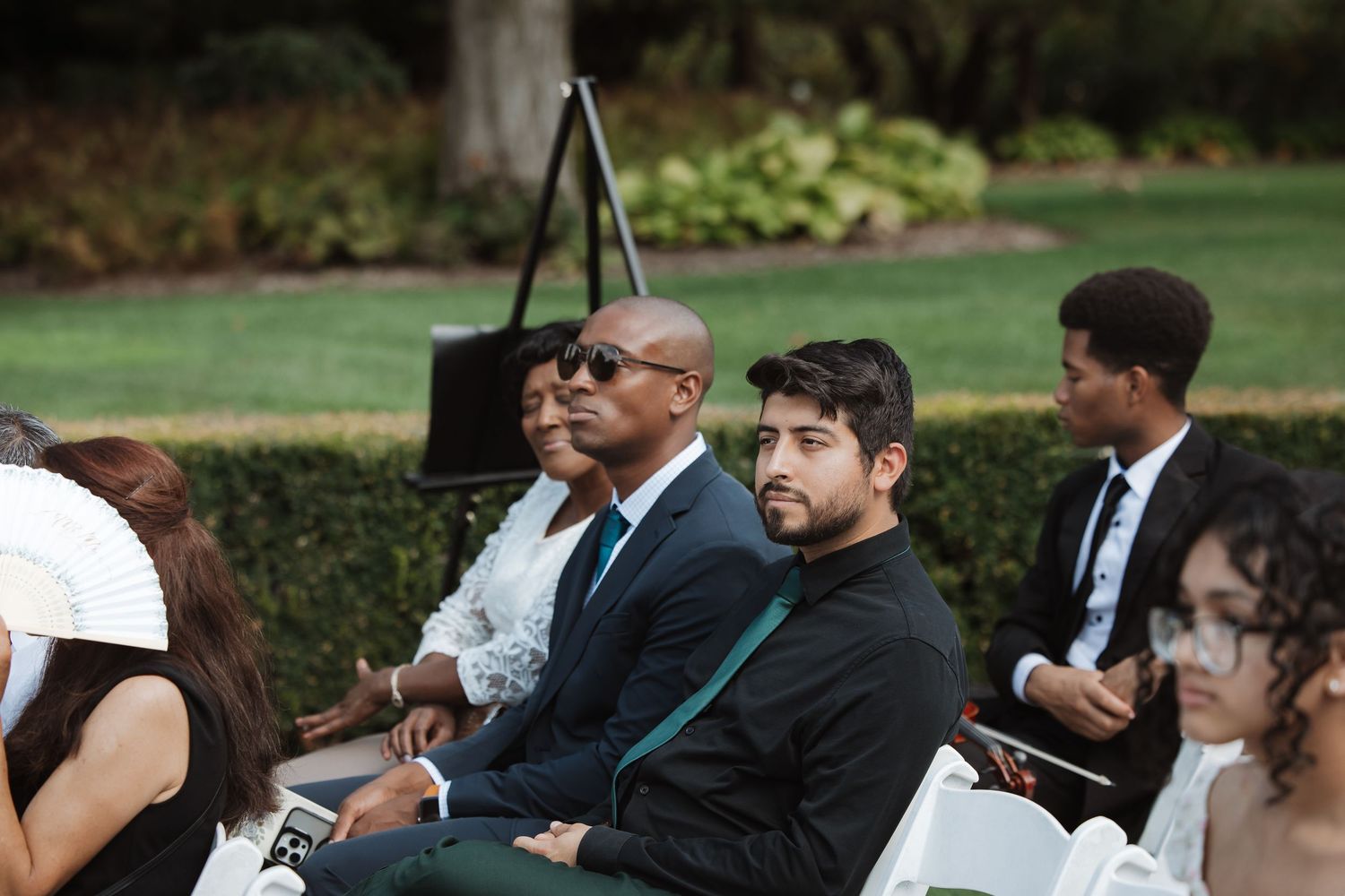 Wedding guests in formal attire seated on white chairs at an outdoor garden ceremony.
