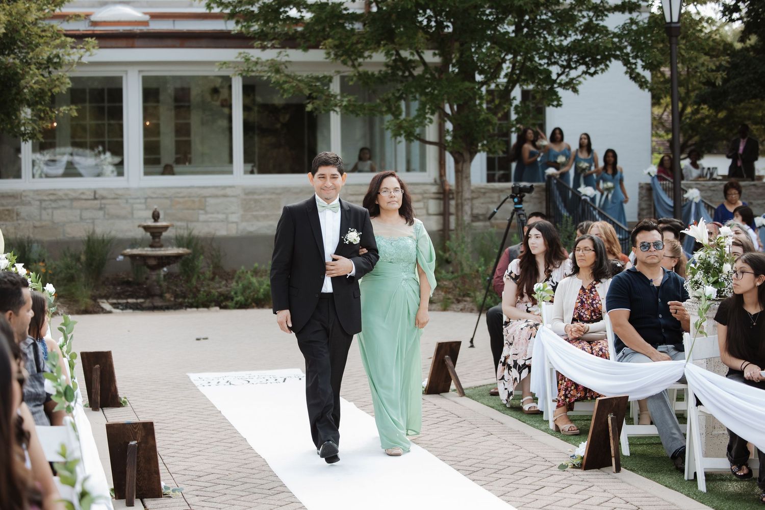 Groom in black tuxedo is escorted down white aisle runner by mother at outdoor wedding.