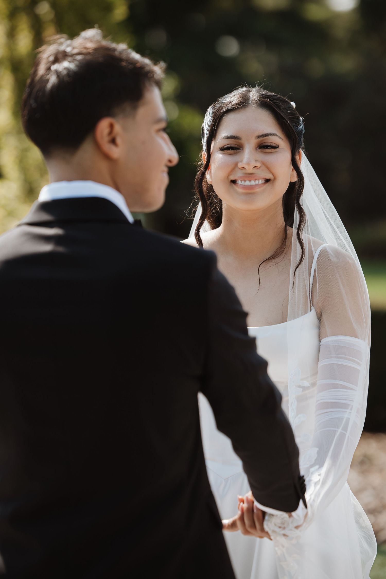 Smiling bride in white veil and dress holds grooms hands during sunny outdoor wedding ceremony.