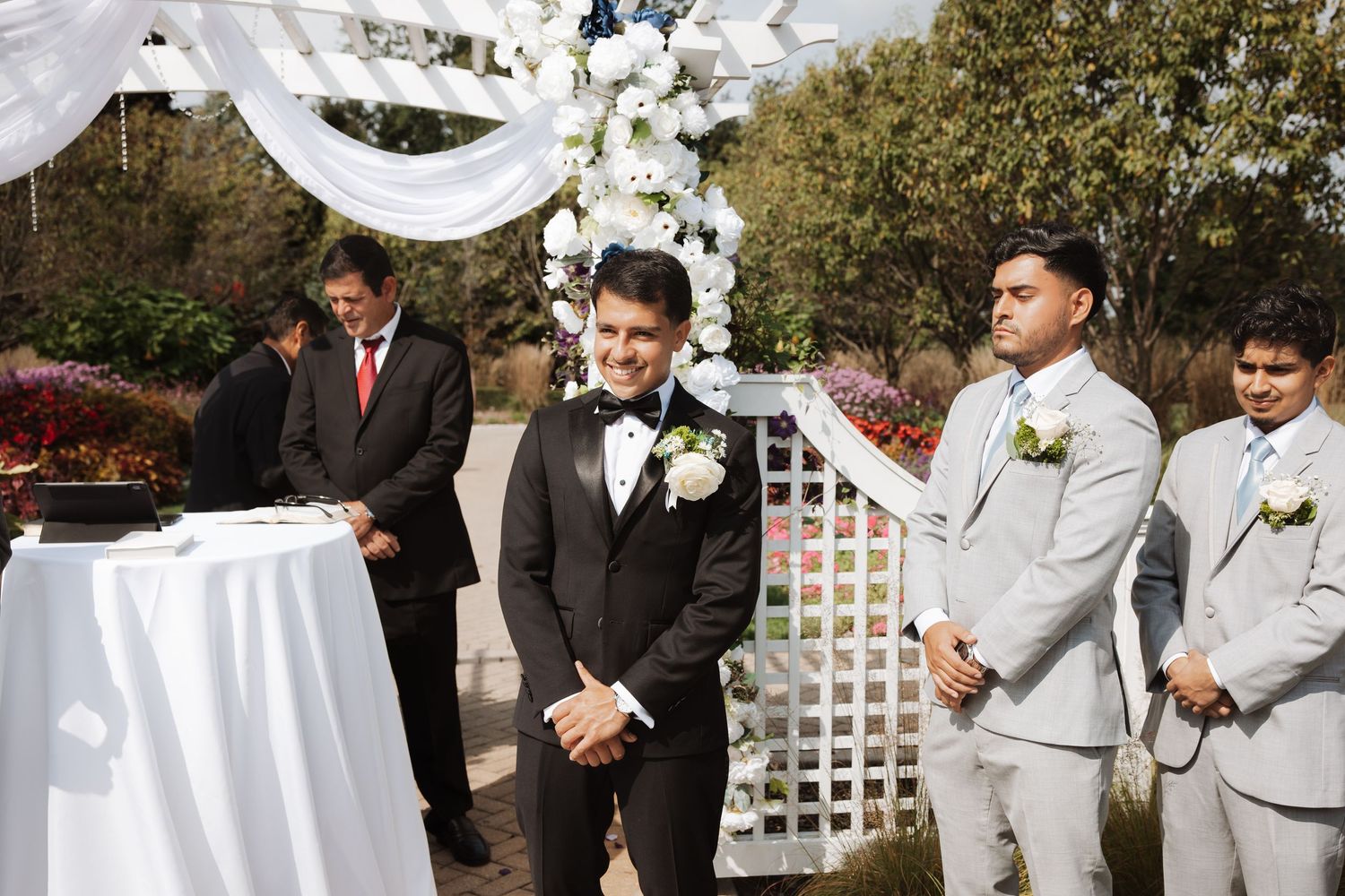 Groom smiling joyfully at outdoor wedding altar flanked by groomsmen in grey suits.